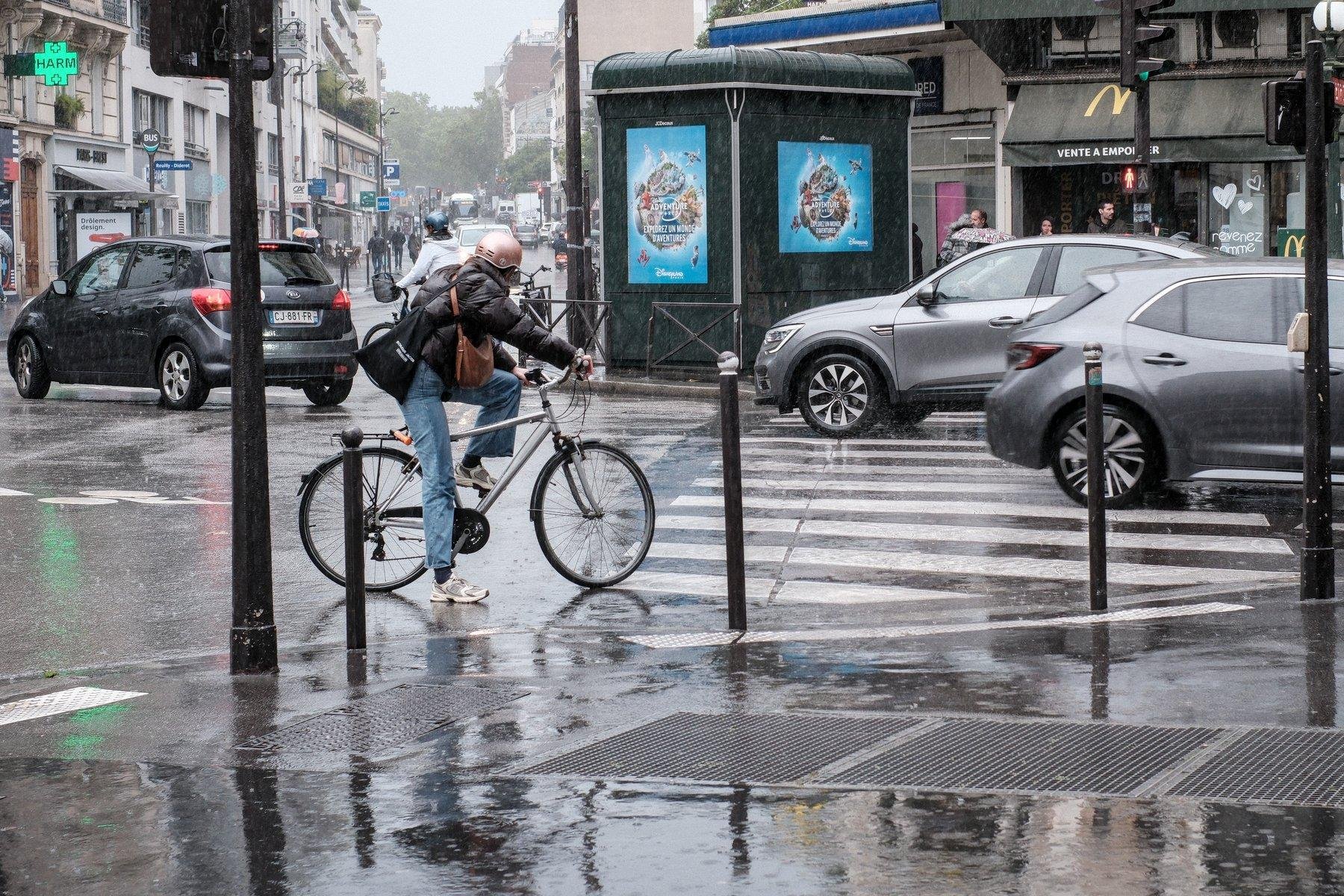 Photo horizontale. Sous la pluie, à un carrefour, une cycliste avec un casque rose, au jean et blouson noir trempé, 2 sacs en bandoulières, attends de pouvoir traverser la route