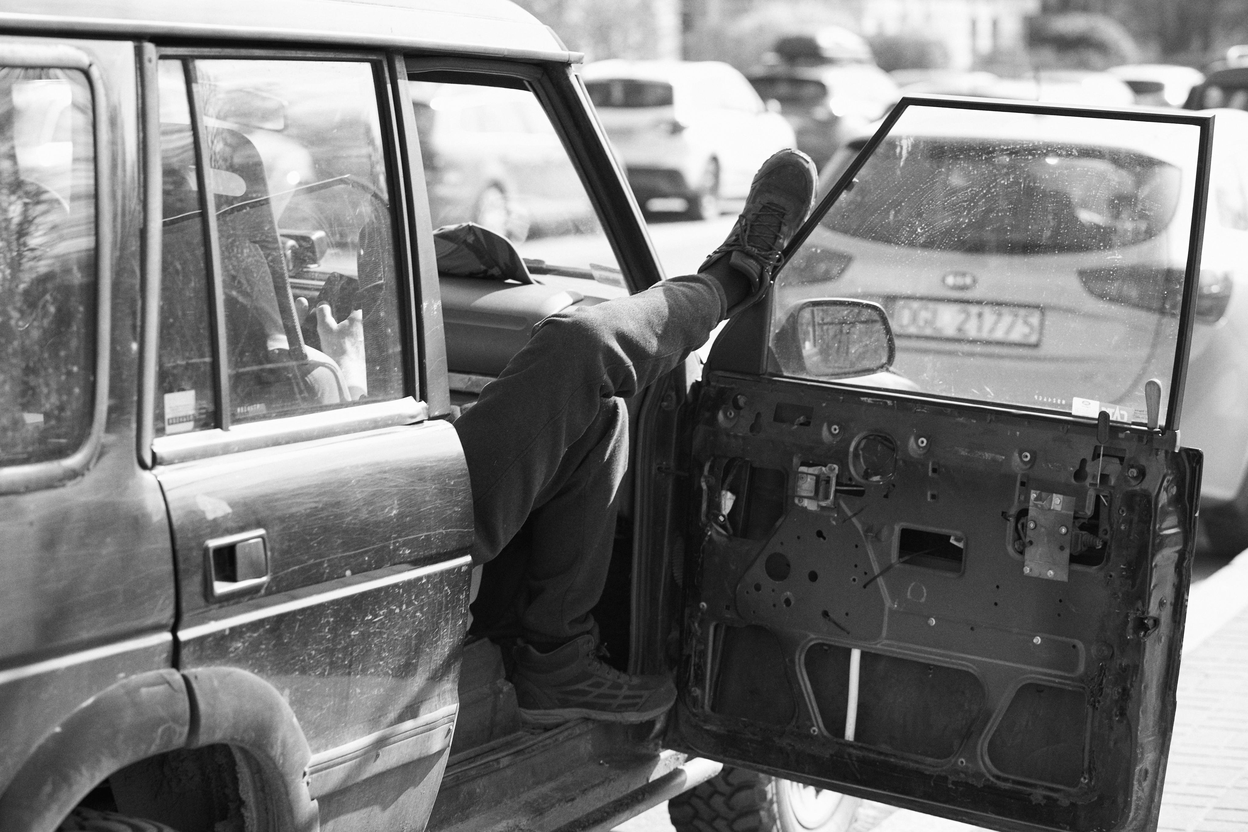 The image shows a person sitting inside a vehicle, with one leg resting on the open front door. The door appears to be missing its interior panel, exposing its mechanical components. Other cars are visible in the background, suggesting a parking area. The photo is in black and white.