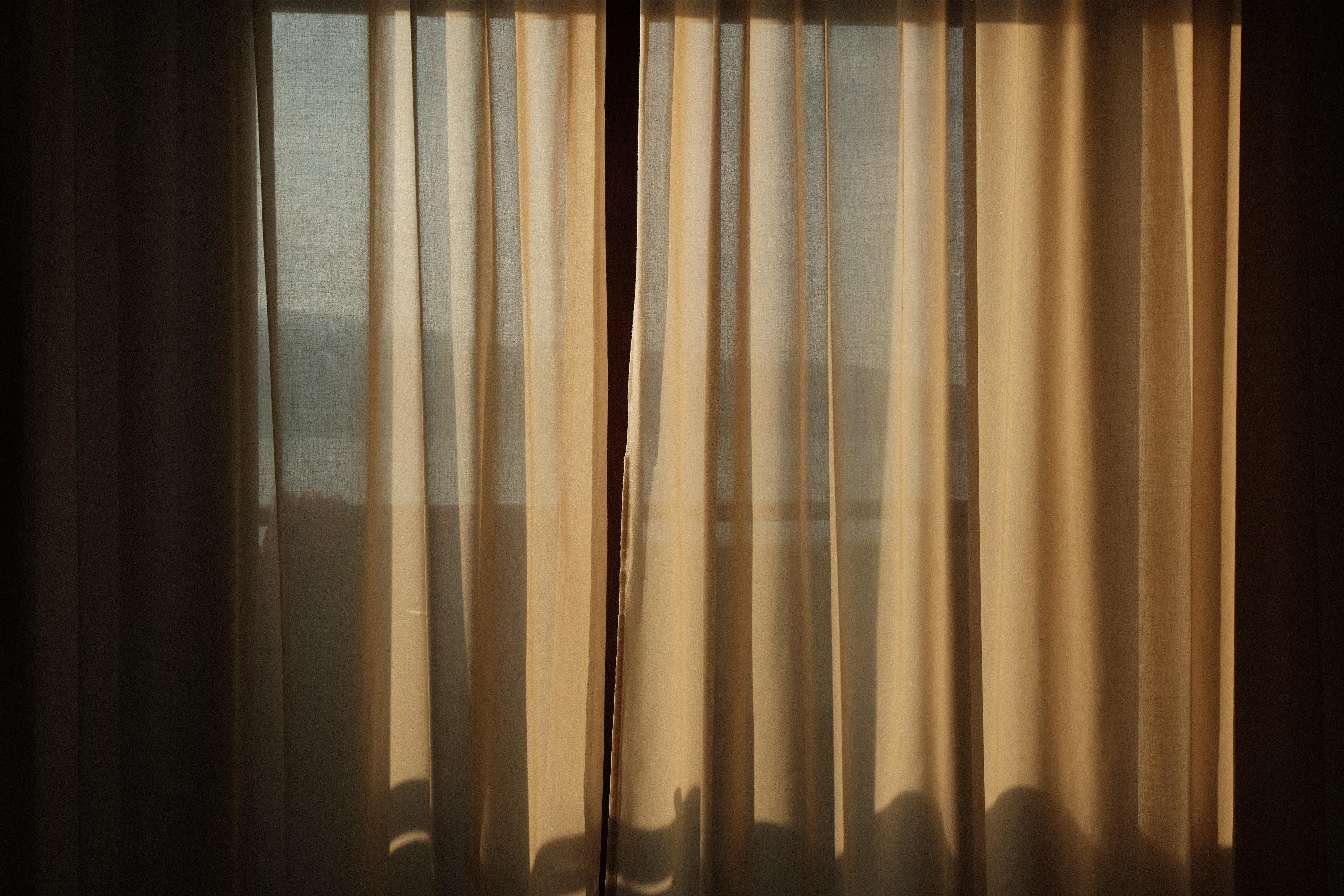 A view of a window lit in early sunlight, the creases of the curtain generating a striped pattern of light and shadow. Through the window you can see a blurry lake and shallow mountain range.