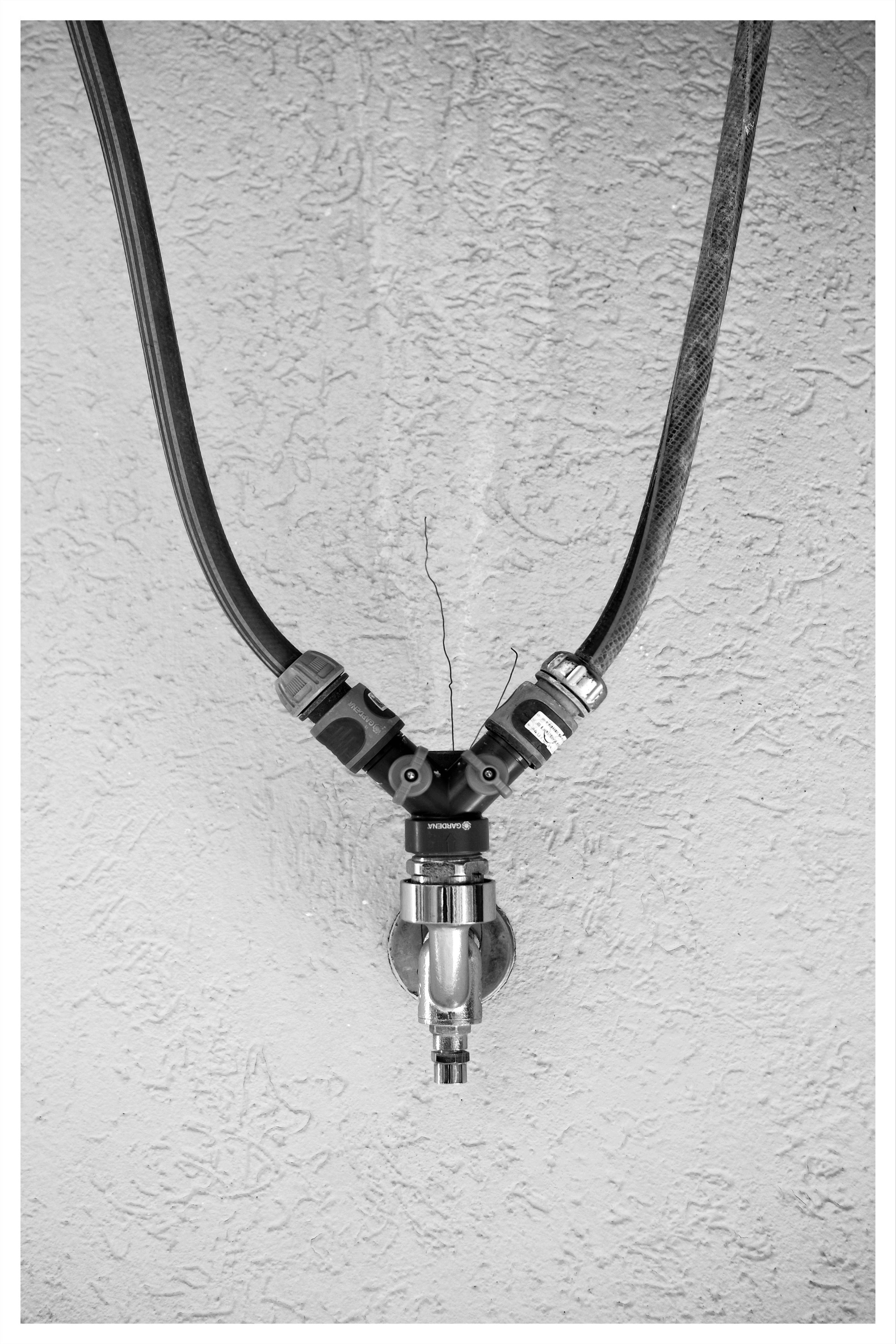 Two black hoses run down from the top corners and join a central Y-shaped manifold attached to a textured wall; a chrome-plated valve and a quick-release coupling hang below it. The picture is upside down.