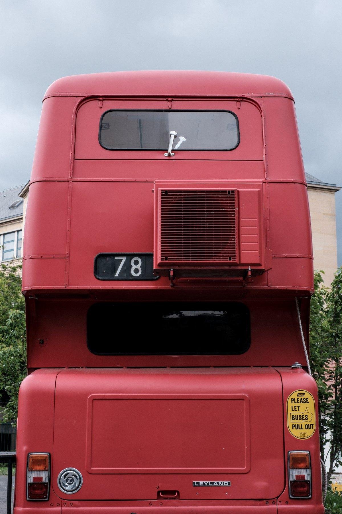 Photo verticale. Vu sur l’arrière d’un bus Londonien rouge à 2 étages. On remarque une unité d’extraction de hotte de cuisine accroché à l’étage, peint du même rouge
