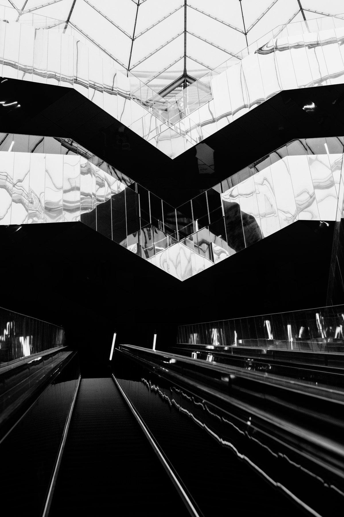 Photo verticale en noir et blanc. Contre plongée depuis un long escalator, vers l’angle d’un passage dont les côté sont en métal réfléchissants. L’ombre du passage et les reflets forment un X