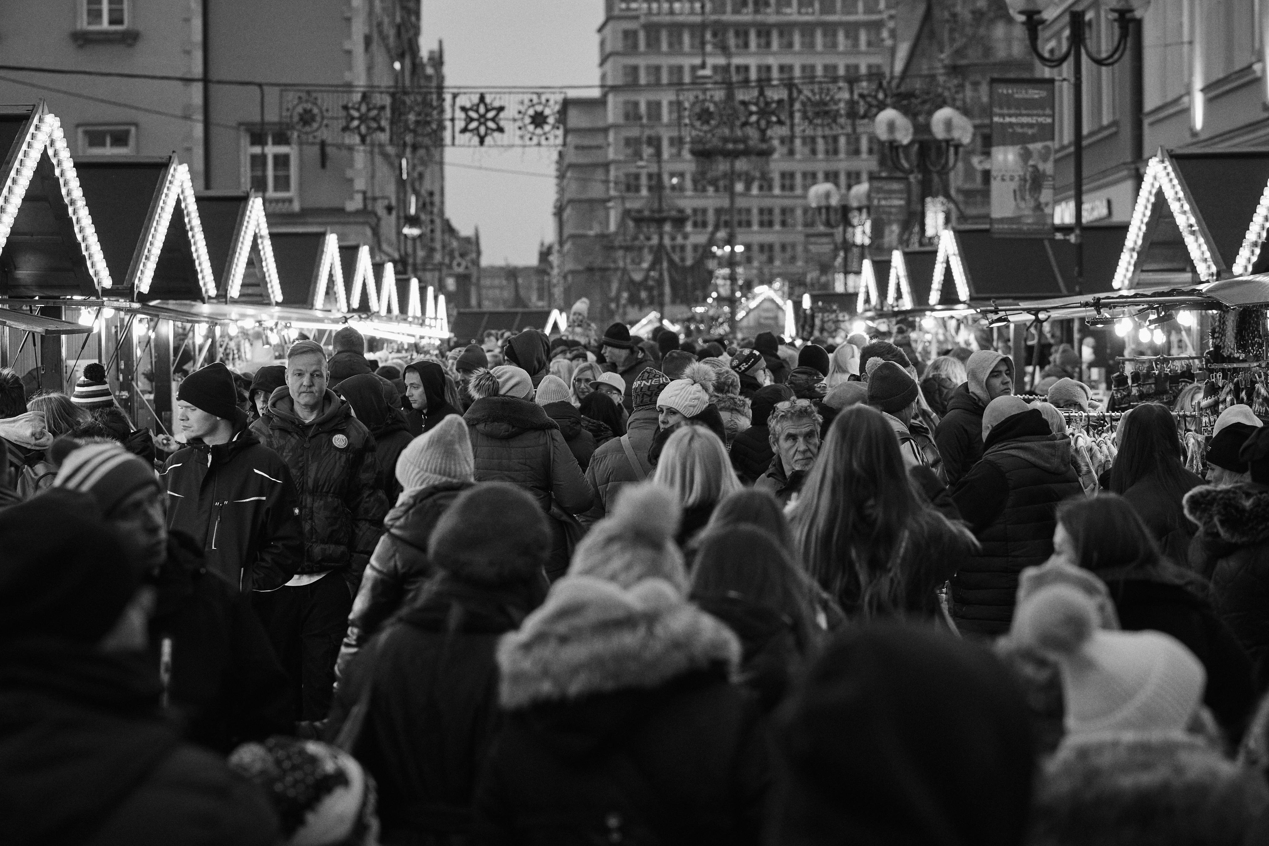 This image shows a crowded outdoor market or street fair, likely during the holiday season given the winter clothing and decorative lights. There are numerous people walking between stalls with peaked roofs, which are lit with strings of lights. The scene is bustling, and the buildings and decorations suggest a festive atmosphere.