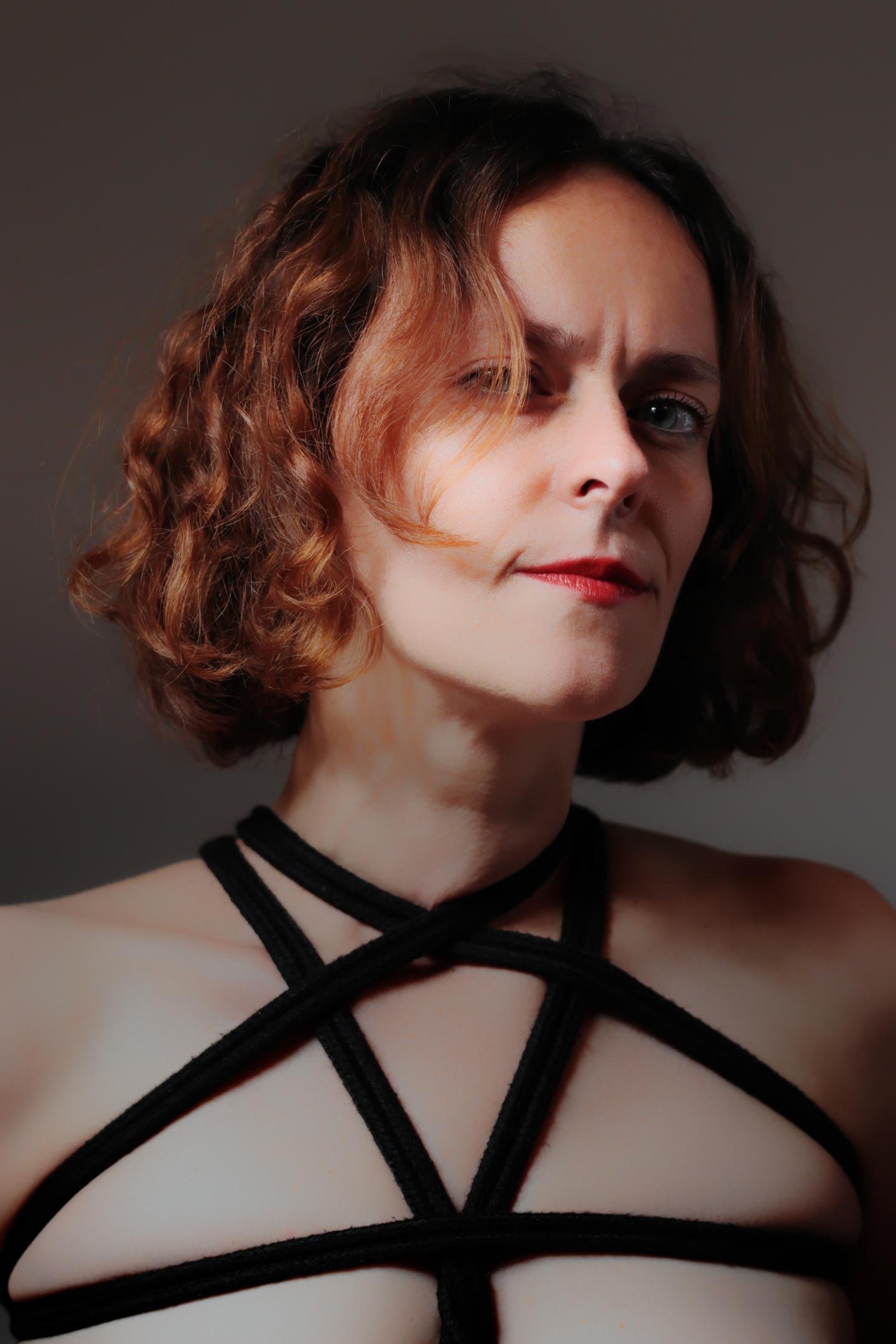 Woman with shoulder-length, curly brown hair wearing a black harness out of rope with crisscrossed straps. The background is neutral, allowing focus on her face and attire. Her expression is neutral, and the lighting highlights her features, creating subtle shadows.