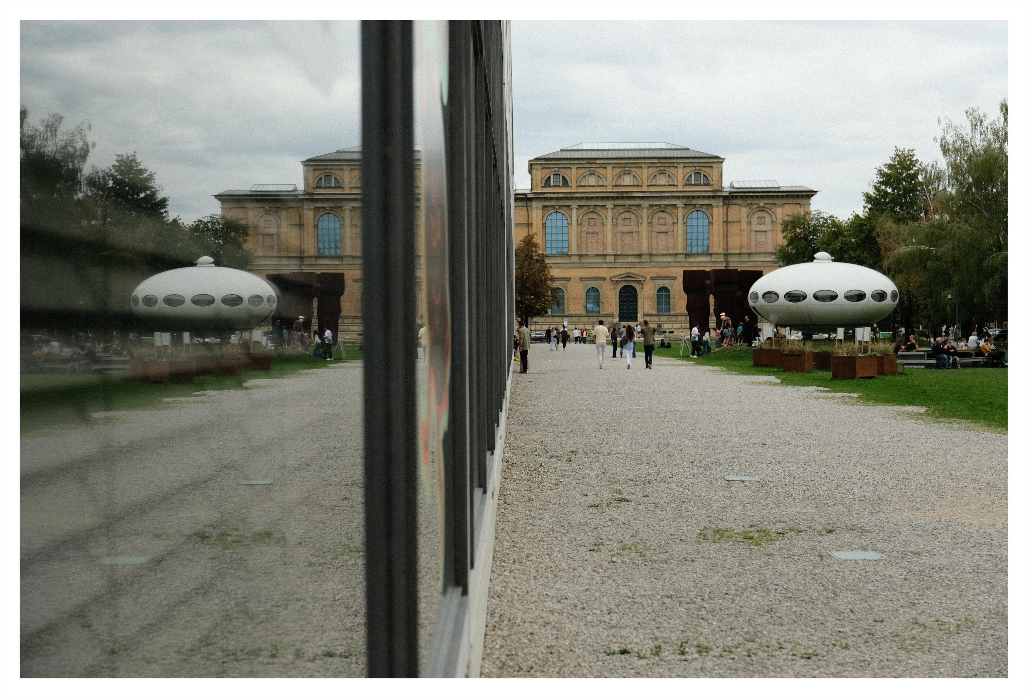 Gravel walkway leading to a large stone building, split by a tall reflective wall down the center; the left side shows blurred reflections while the right side shows people walking and a white oval pavilion structure on the lawn under a cloudy sky.
