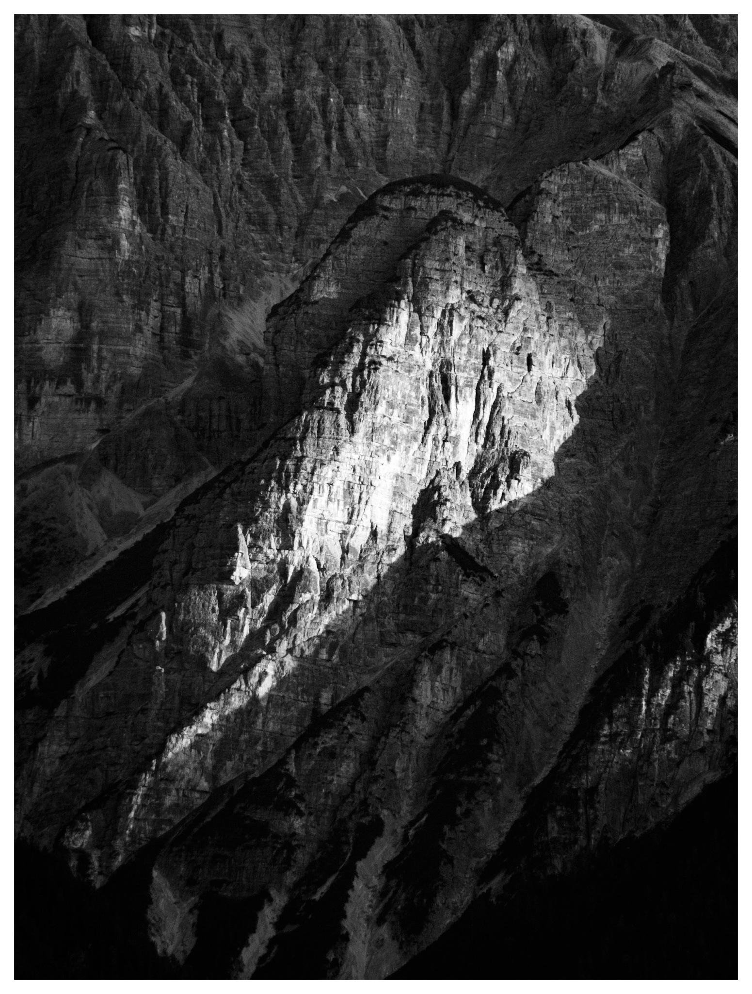 Black-and-white close-up of a rugged mountain face, with a bright sunlit rock ridge centered against darker shadowed cliffs and deep crevices.