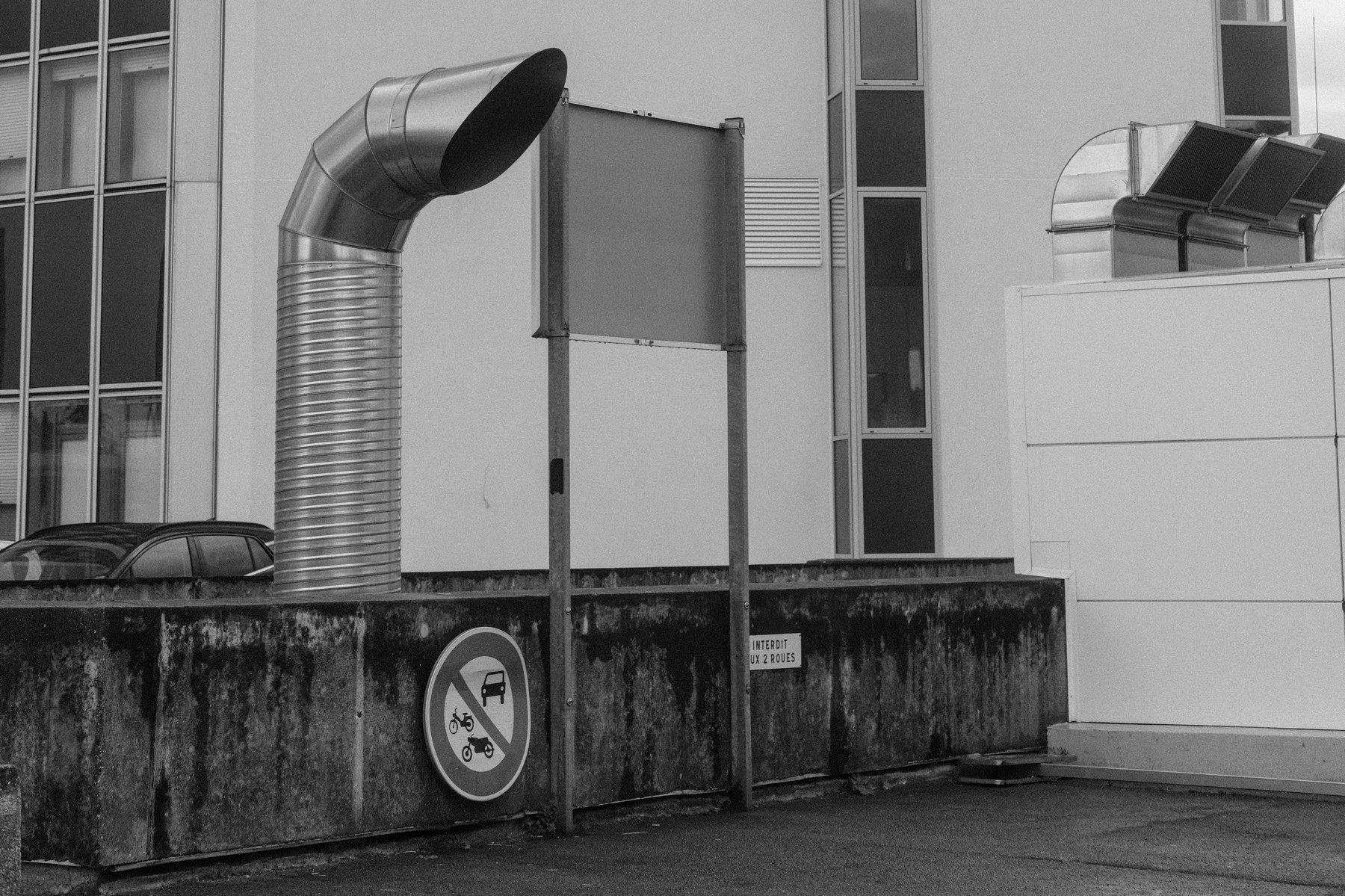 Photo horizontale en noir et blanc. Un gros tuyau d’évacuation en métal texturé comme une vis sort d’entre 2 muret de sécurité et se fini comme un périscope