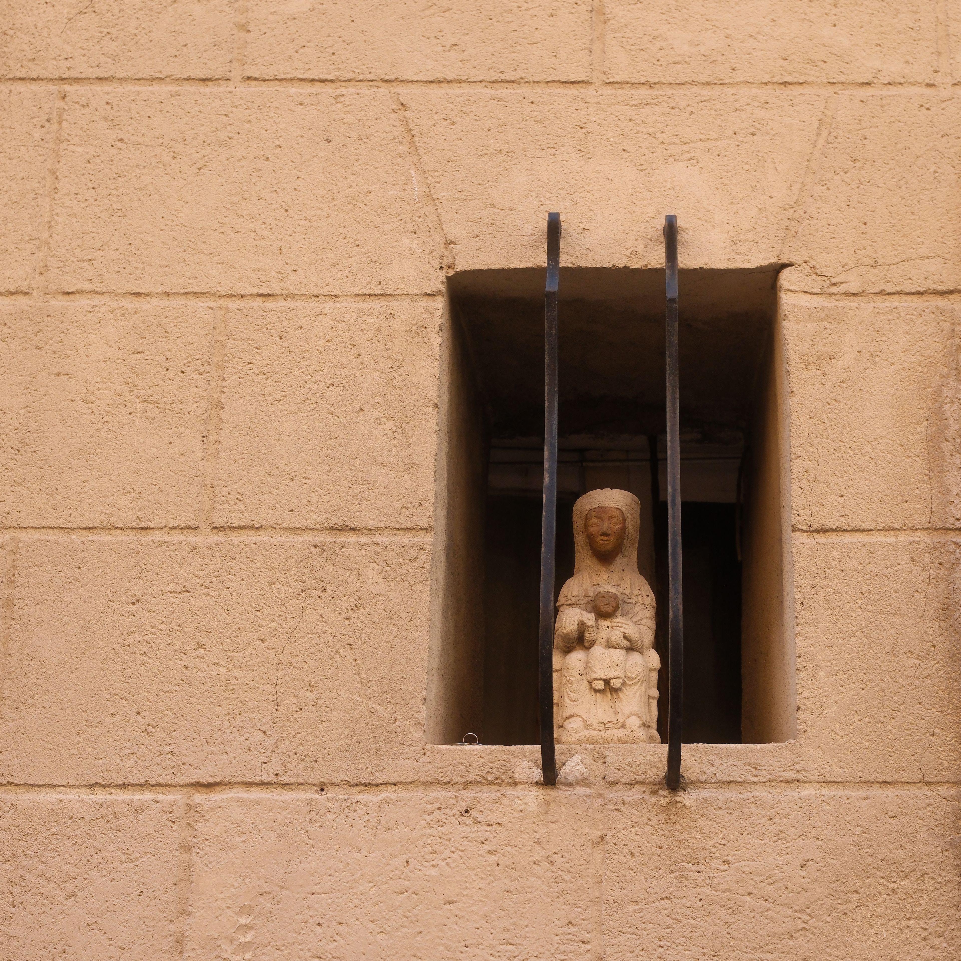 Photo format carré, montrant, décentré à droite une alcove rectangulaire dans un mur couleur orange pâle avec deux barreaux verticaux, protégeant une statuette de femme tenant un enfant sur ses genoux