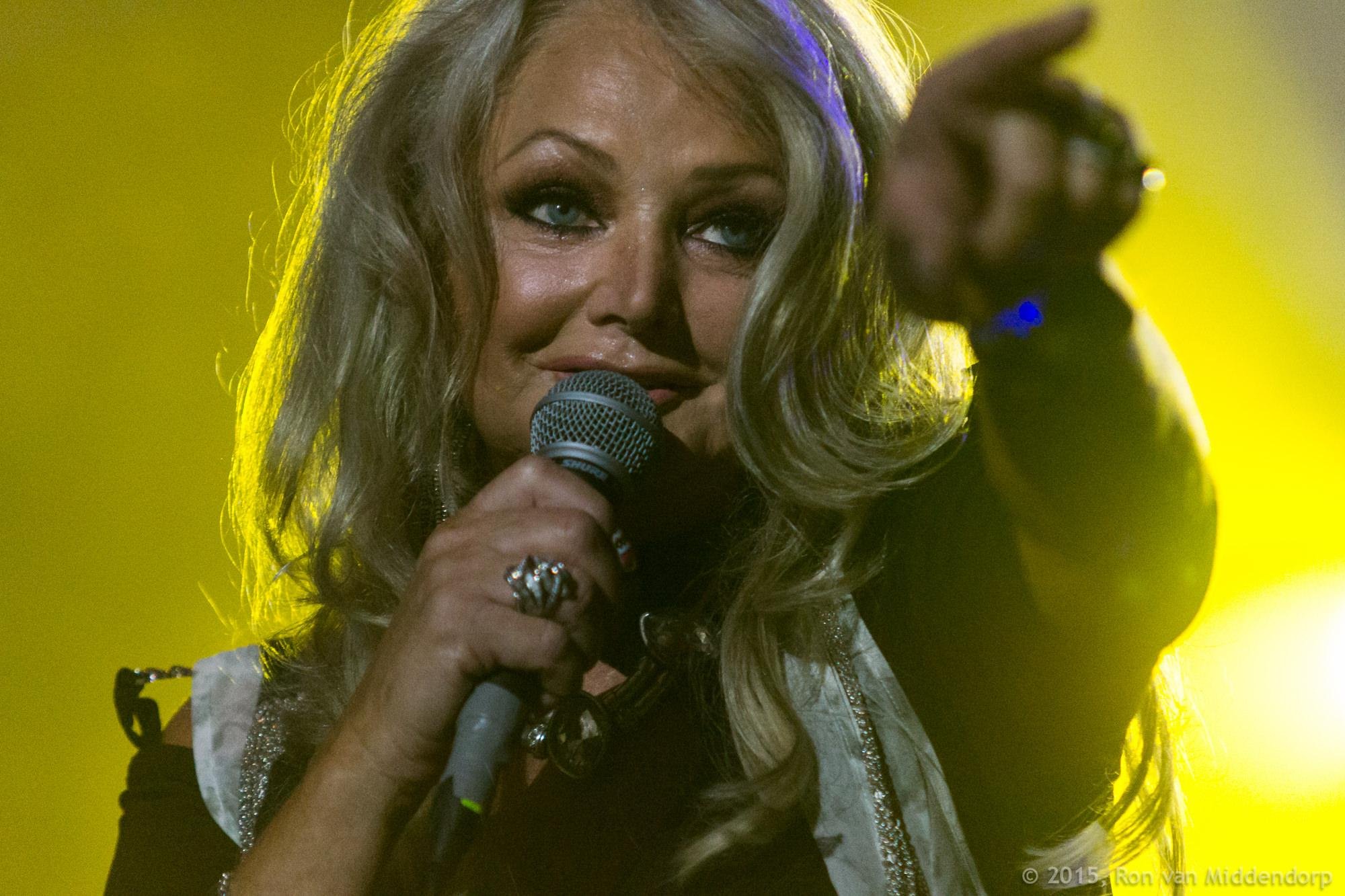 Close-up of Bonnie Tyler on stage holding a microphone near her mouth and pointing toward the audience, with bright yellow stage lighting in the background.