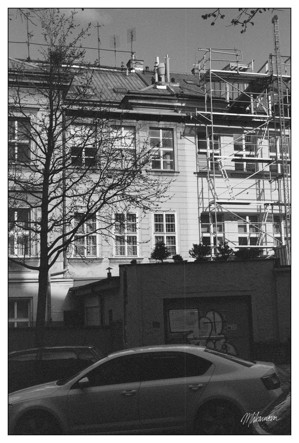 Building with scaffolding on the right side, partially obscured by a leafless tree in the foreground. The structure features multiple windows and a sloped roof with various antennas and chimneys. A parked car is visible at the bottom, with graffiti on the garage door behind it. The image is in black and white.