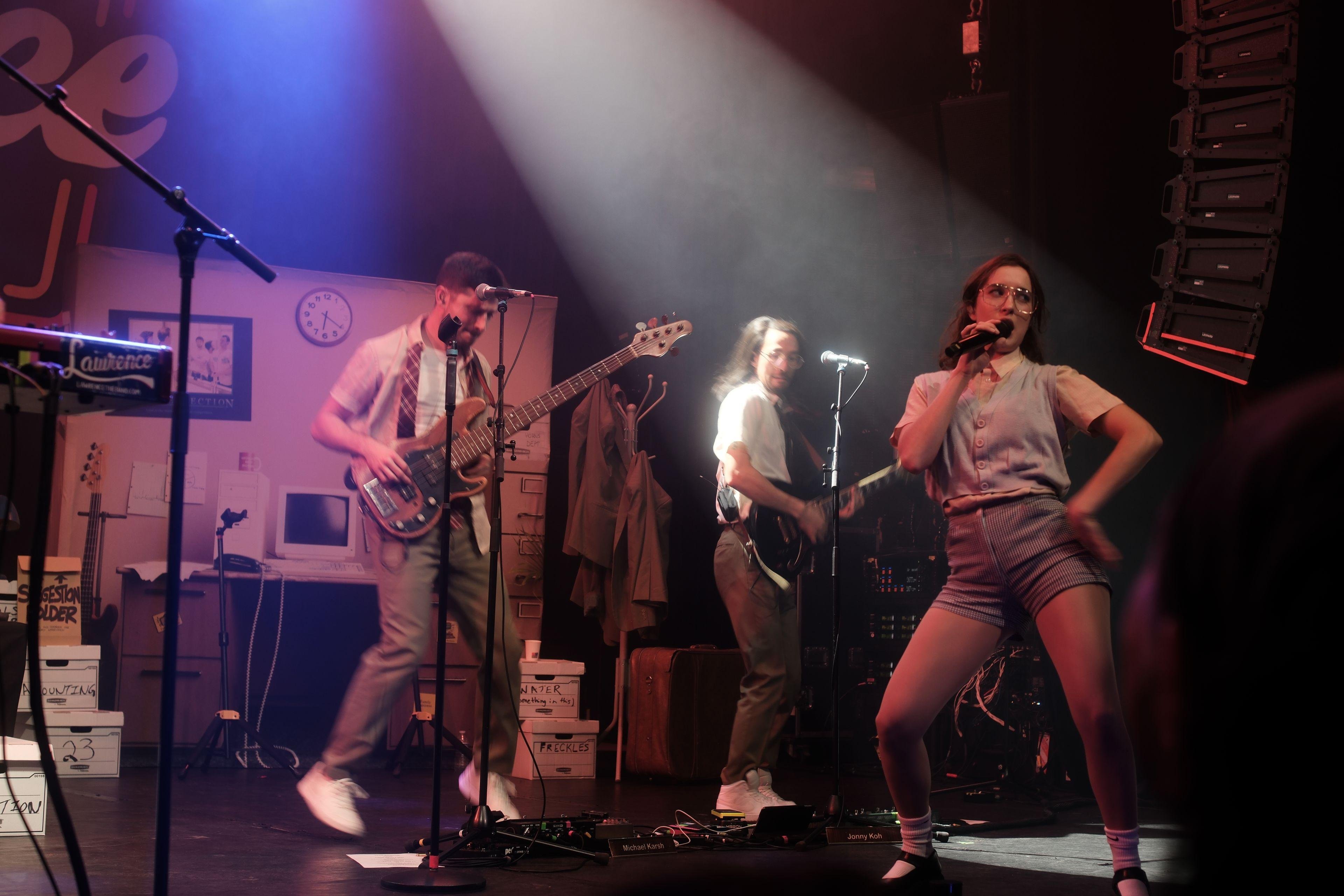 Photo horizontale. Au 1er plan, Gracie Lawrence entrain de danser et chanter, le dos de sa main gauche appuyé sur sa hanche, la main droite tenant le micro. En arrière plan, le bassiste et le guitariste dansent en synchro