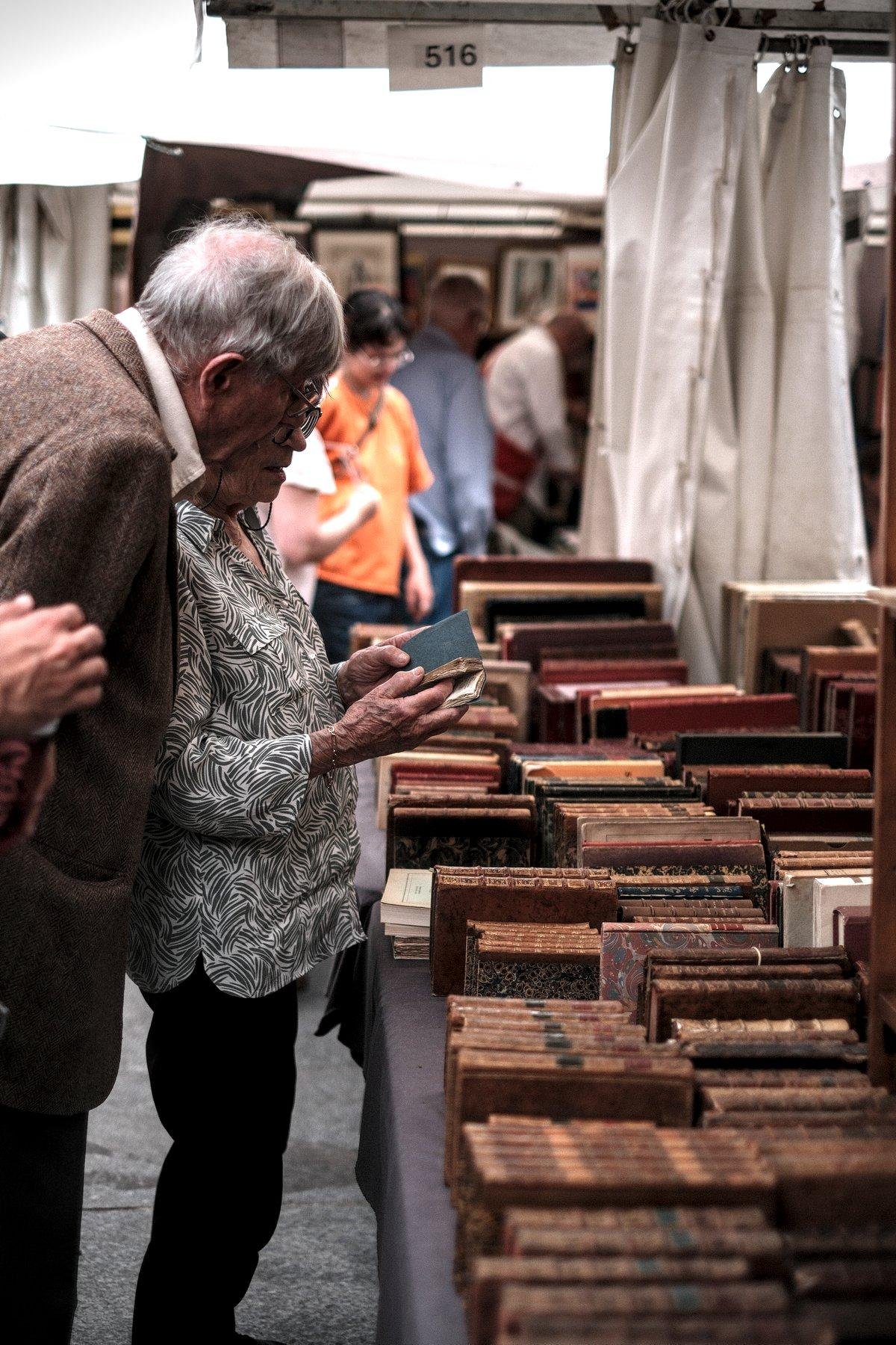 Photo verticale. Devant un étalage plein de livres anciens reliés cuir, une dame en chemise blanche à motifs abstraits noir montre un petit livre à son mari au cheveux blancs, veste marron qui se penche un peu par dessus son épaule
