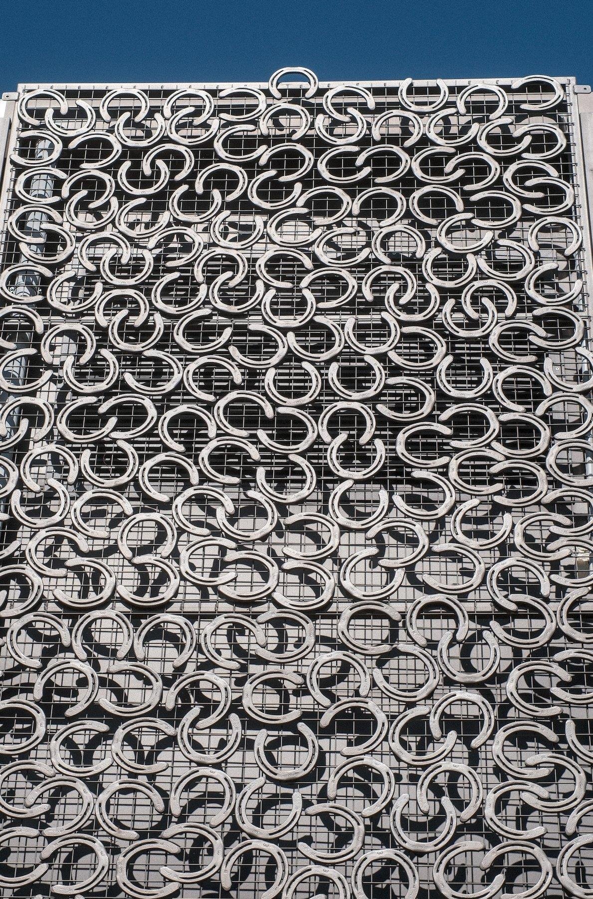 Photo verticale. Sur la façade d’un bâtiment, une grille est fixée sur le mur, sur laquelle des dizaine de fers à cheval gris métal sont accrochés dans tous les sens. Sous un ciel bleu