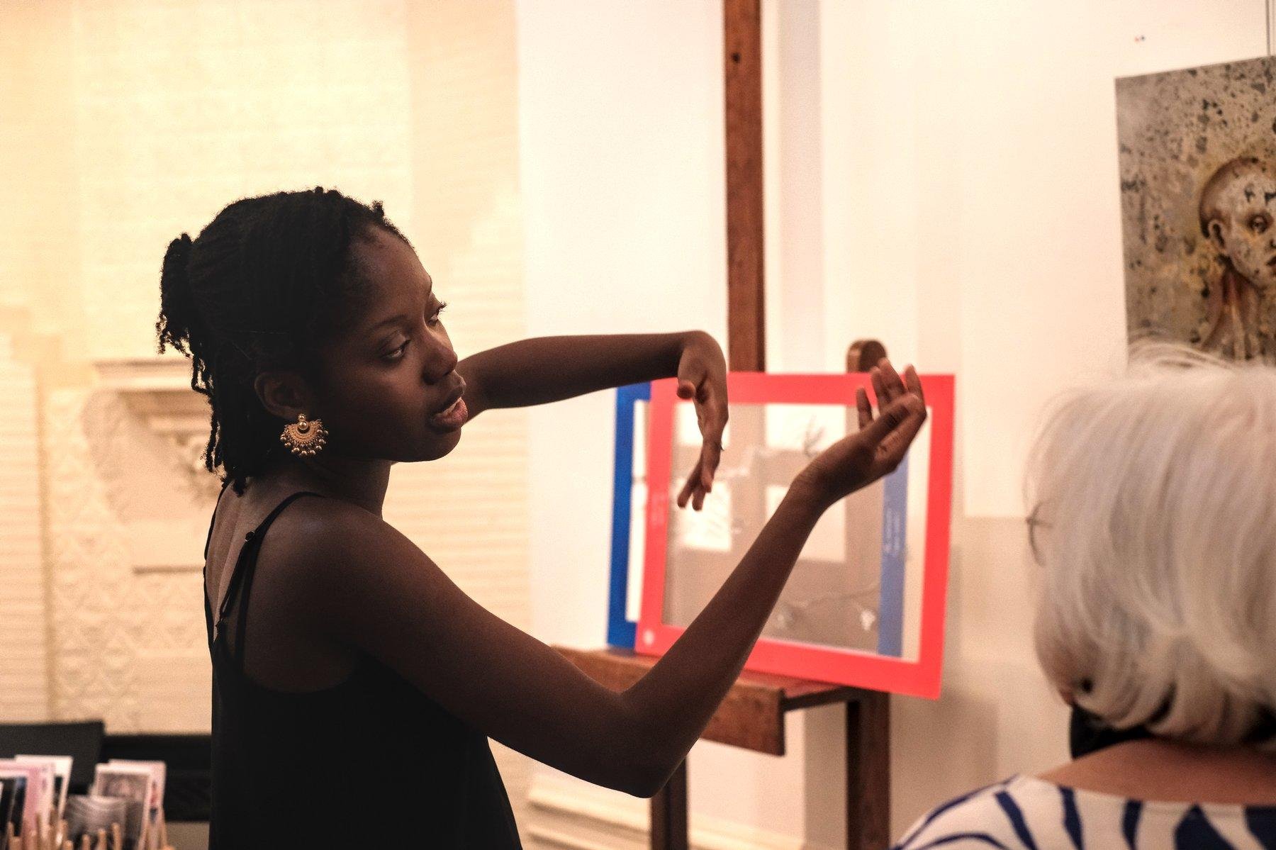 Photo horizontale. Une femme aux cheveux noir en dreadlocks rassemblés en chignon, en débardeur noir, fait des gestes en parlant à une dame aux cheveux blanc. On aperçoit un bout de toile de peinture et devine qu’elle explique son œuvre