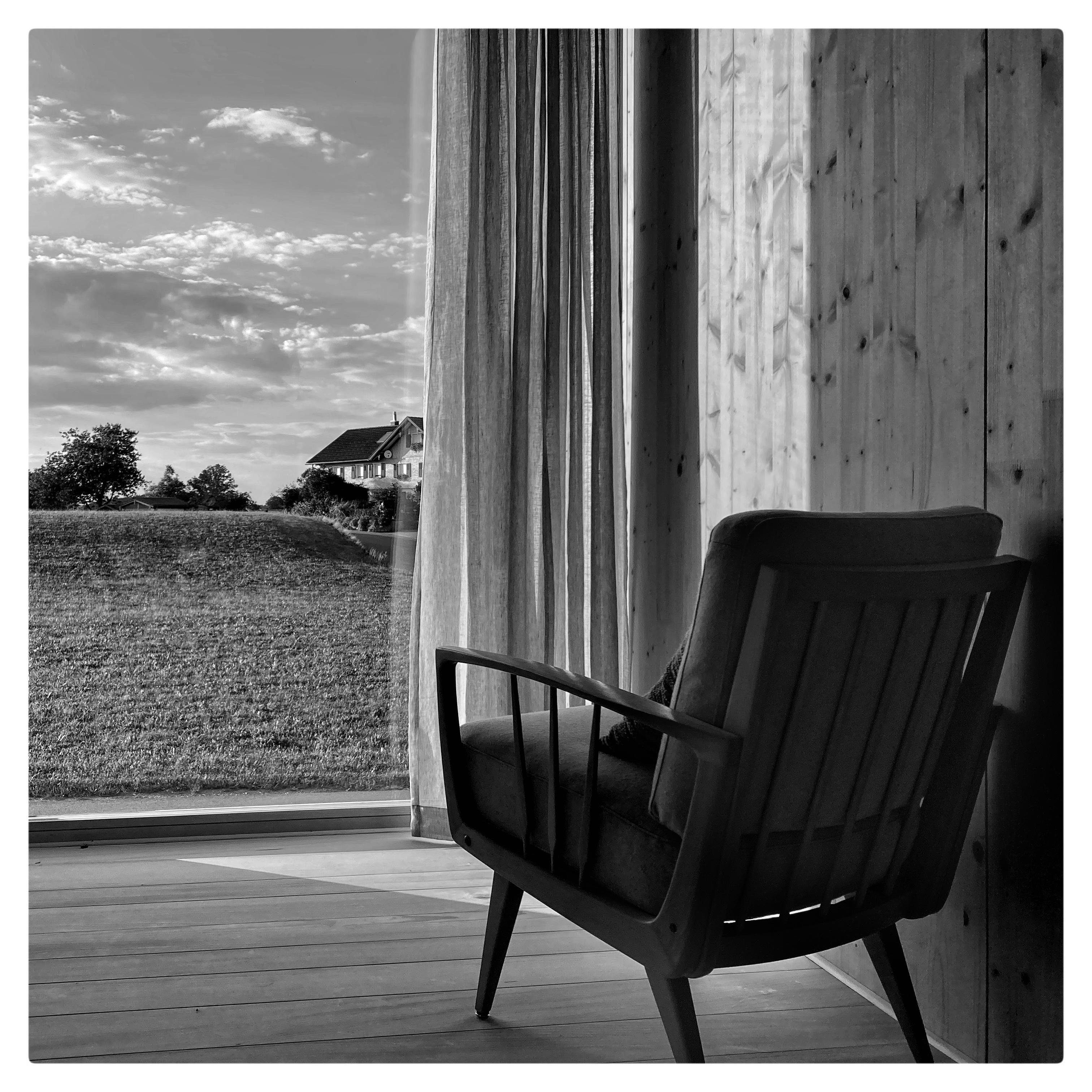 Black and white image of an empty wooden armchair facing a floor-to-ceiling window with sheer curtains, overlooking a grassy field and house under a cloudy sky. Sunlight casts shadows on the wooden floor and wall.