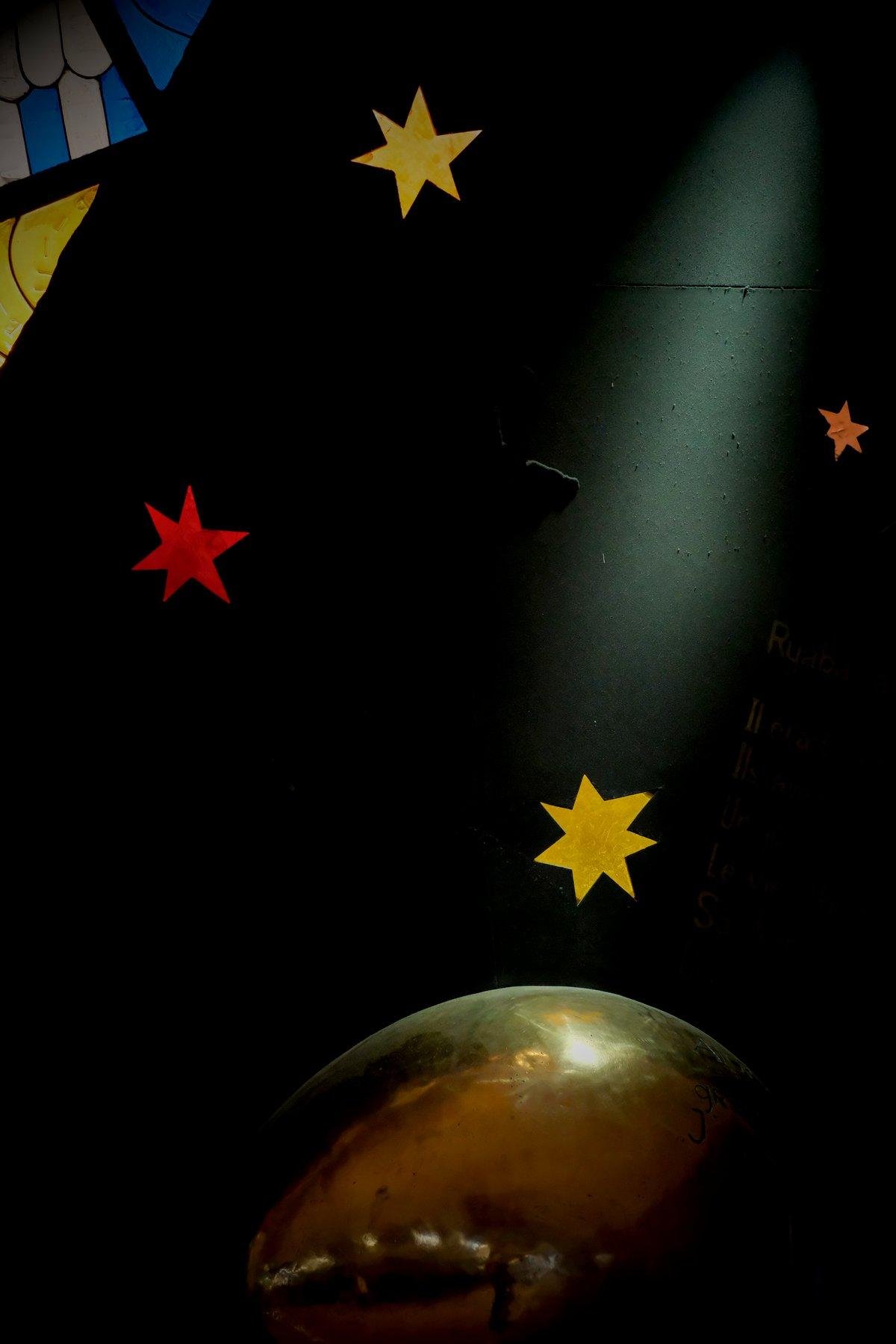 Photo verticale. Sur un mur noir, une œuvre artistiques. Des étoiles comme en papier collées sur le mur, et en bas, une sphère dorée en métal poli représentant le soleil je suppose. Un spot vient éclairer la sphère.