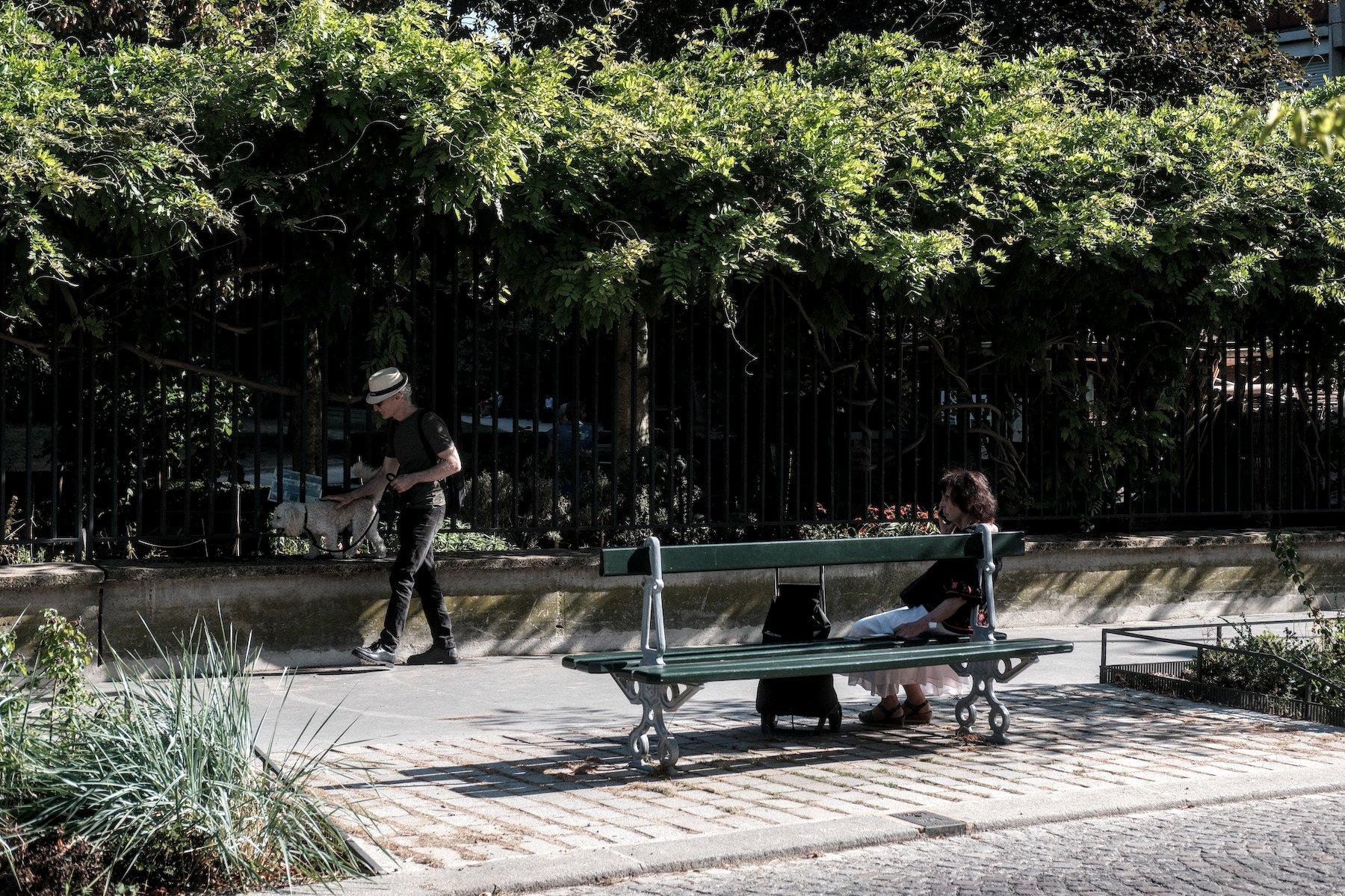 Photo horizontale. À droite, une dame assise sur un double banc publique parisien vert, côté trottoir, face à une grille en fer noir sous des arbres. À gauche, le long de cette grille, un homme au chapeau blanc, t-shirt noir et jean noir, promène son chien blanc qui trottine sur le muret de la base de la grille