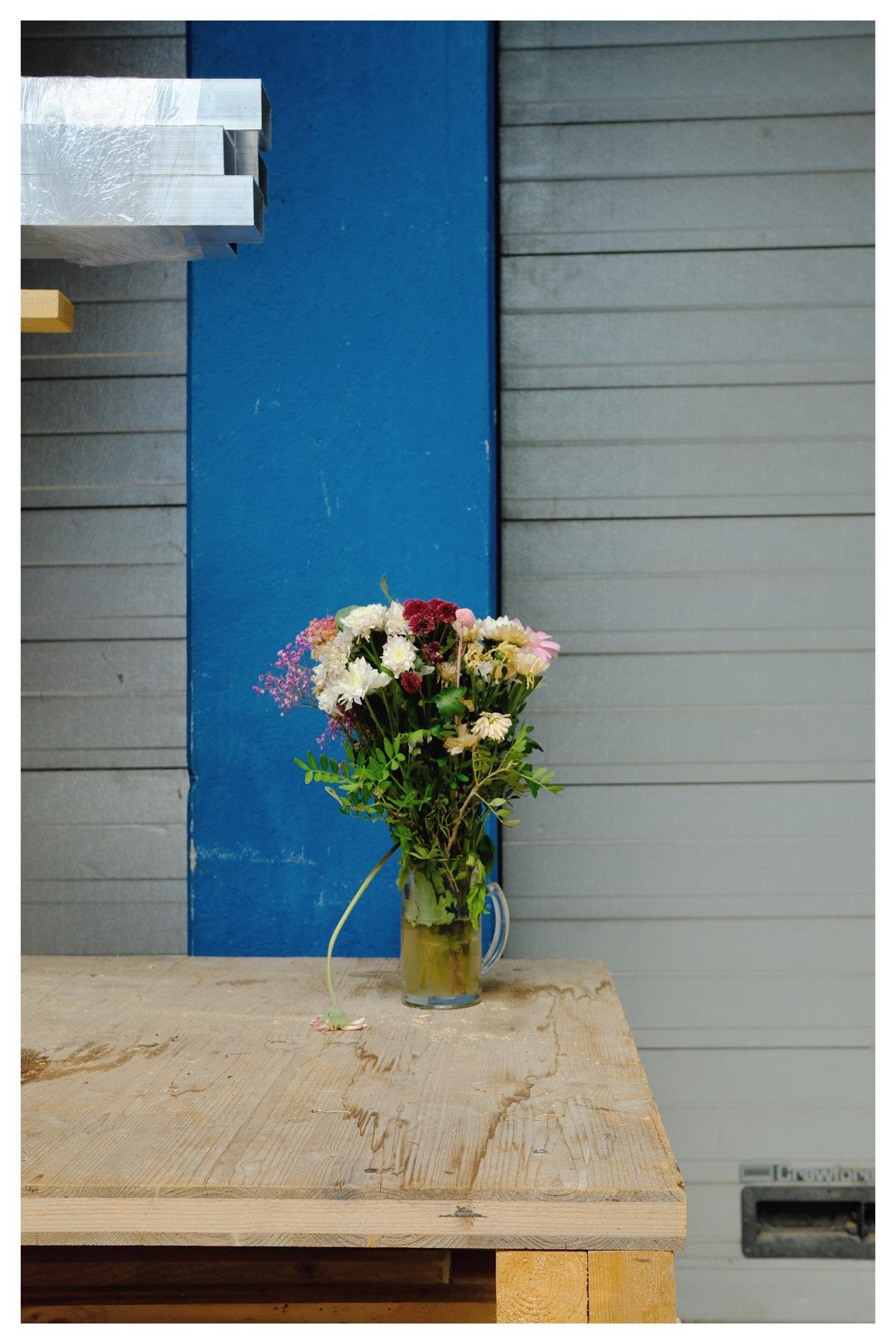 A bouquet of various flowers, including pink, red, and white, is arranged in a glass vase sitting on a wooden table. The background features a vertical blue strip and horizontal gray panels. A wrapped package is partially visible at the top left corner.