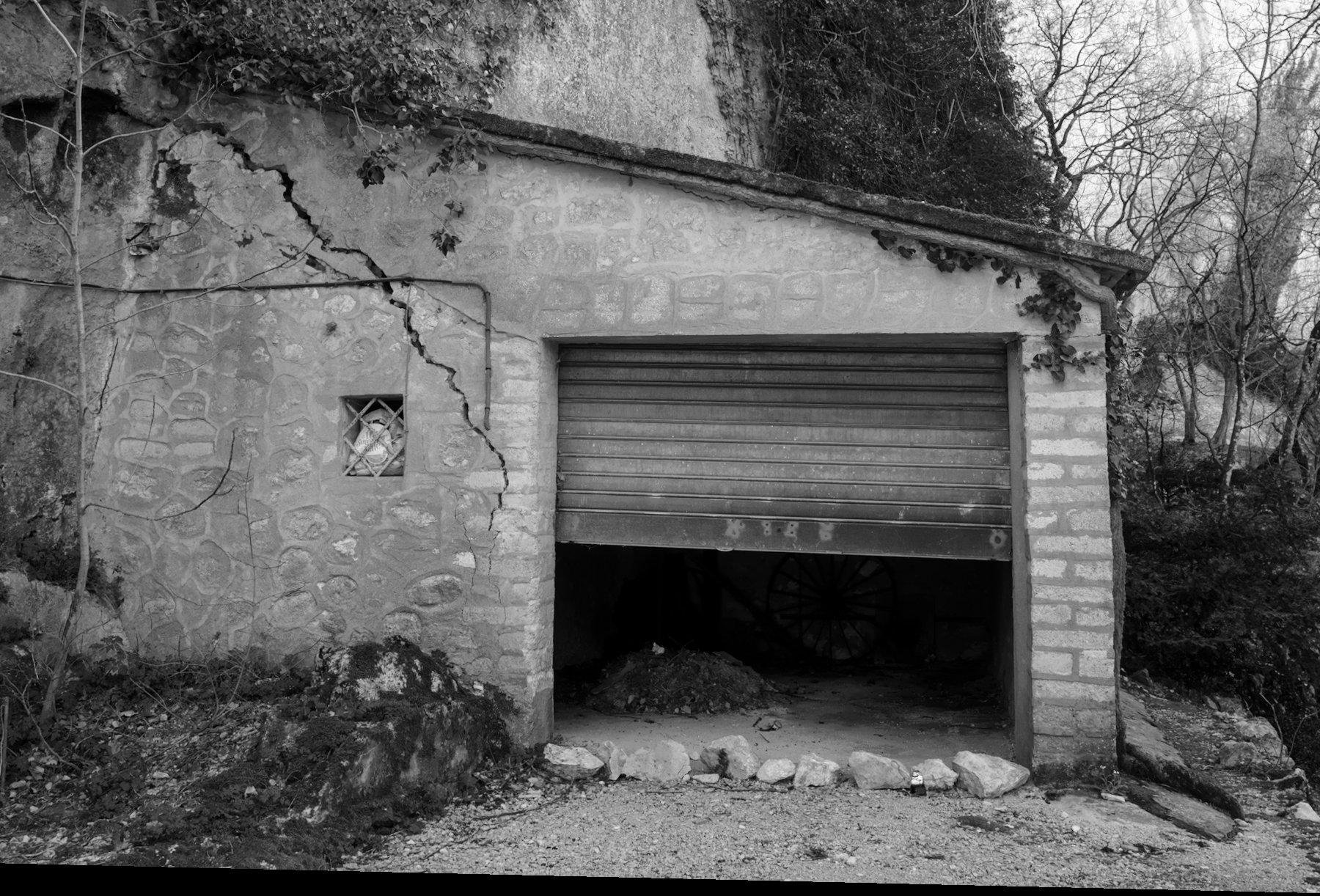 Photo horizontale en noir et blanc. Un garage construit à flanc de montagle, la porte à moitiée ouverte. Une énorme fissure est apparente sur le mur à gauche