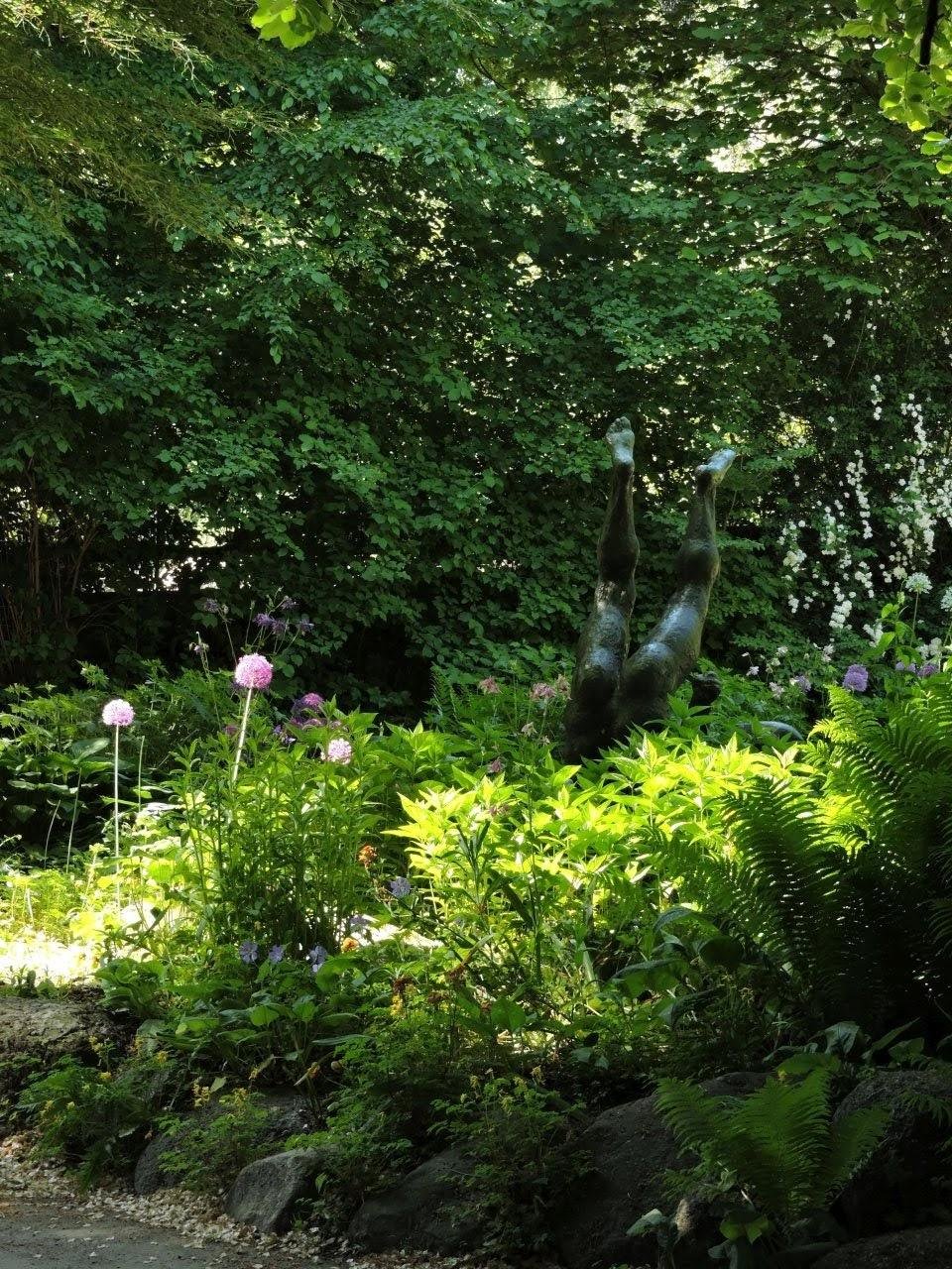 A lush garden with vibrant green foliage, featuring a sculpture of legs pointing upwards. Bright pink and purple flowers, as well as dense ferns and leafy plants, surround the artwork. Sunlight filters through the tree canopy, creating dappled light and shadow effects on the ground.