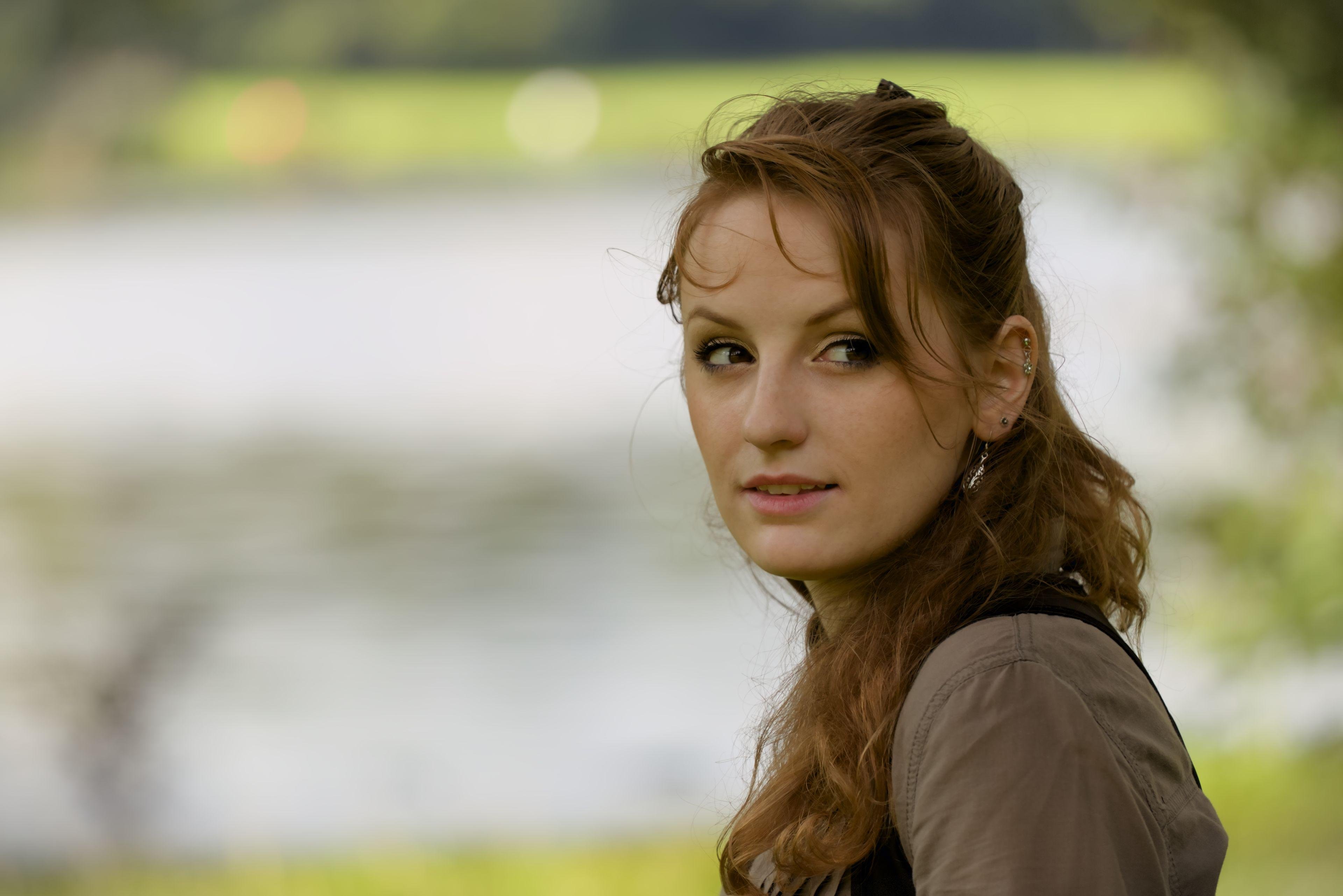 Photo portrait, montrant une jeune femme rousse au cheveux long élégamment coiffé, portant un chemisier marron et par dessus, un corset noir. Elle regarde sur le côté du coin des yeux et en tournant la tête, comme si elle avait entendu du bruit