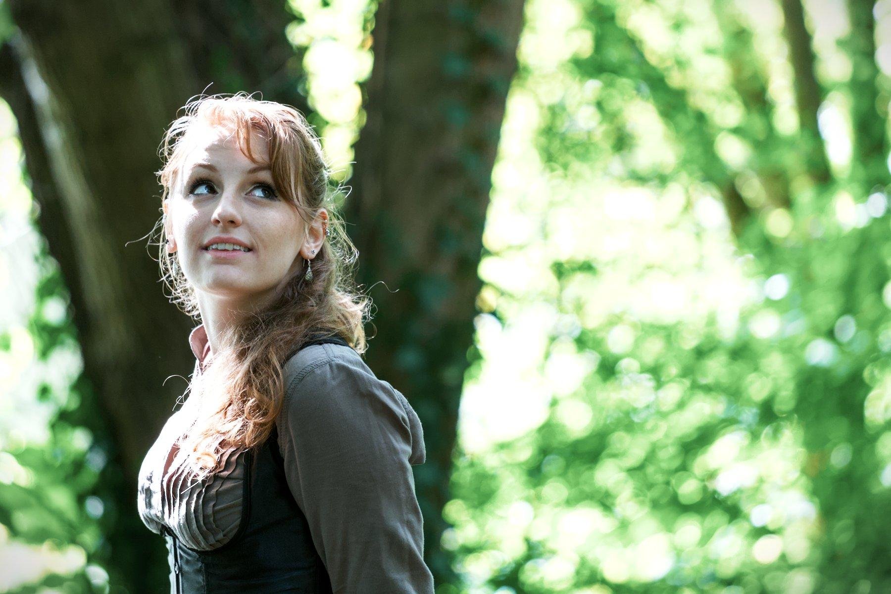 Photo horizontale. Une jeune femme rousse au cheveux long élégamment coiffé, portant un chemisier marron et par dessus, un corset noir, une longue jupe marron foncé. Elle se tiens debout, les mains derrière le dos, regardand en l’air en souriant. Le fond est composé d’un gros arbres et de feuillages verts
