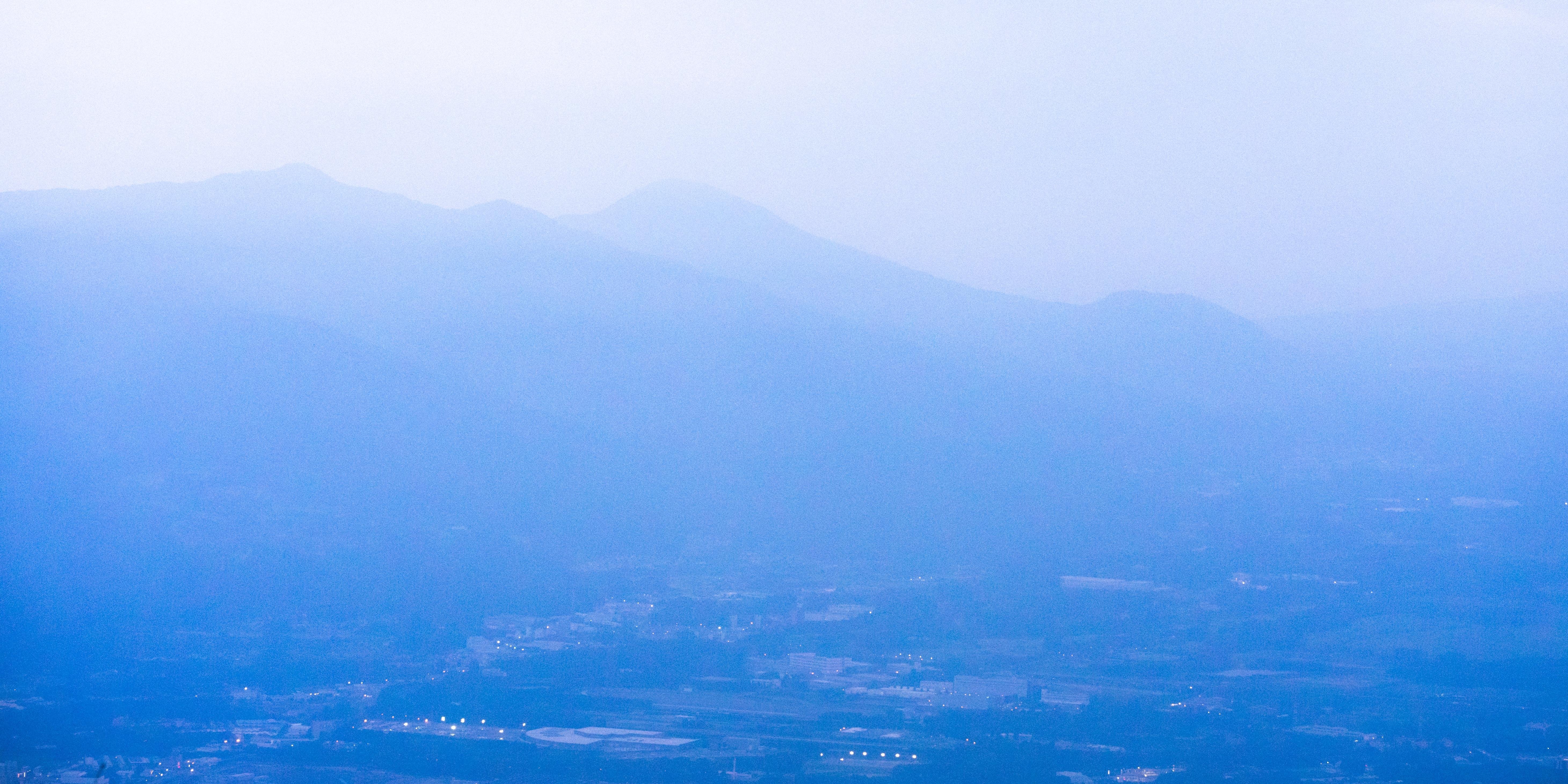 Blue-tinted landscape with layers of distant mountains under a hazy sky. The foreground features a faint view of a valley with scattered buildings and glowing lights, while the mountains gradually fade into fog in the background.