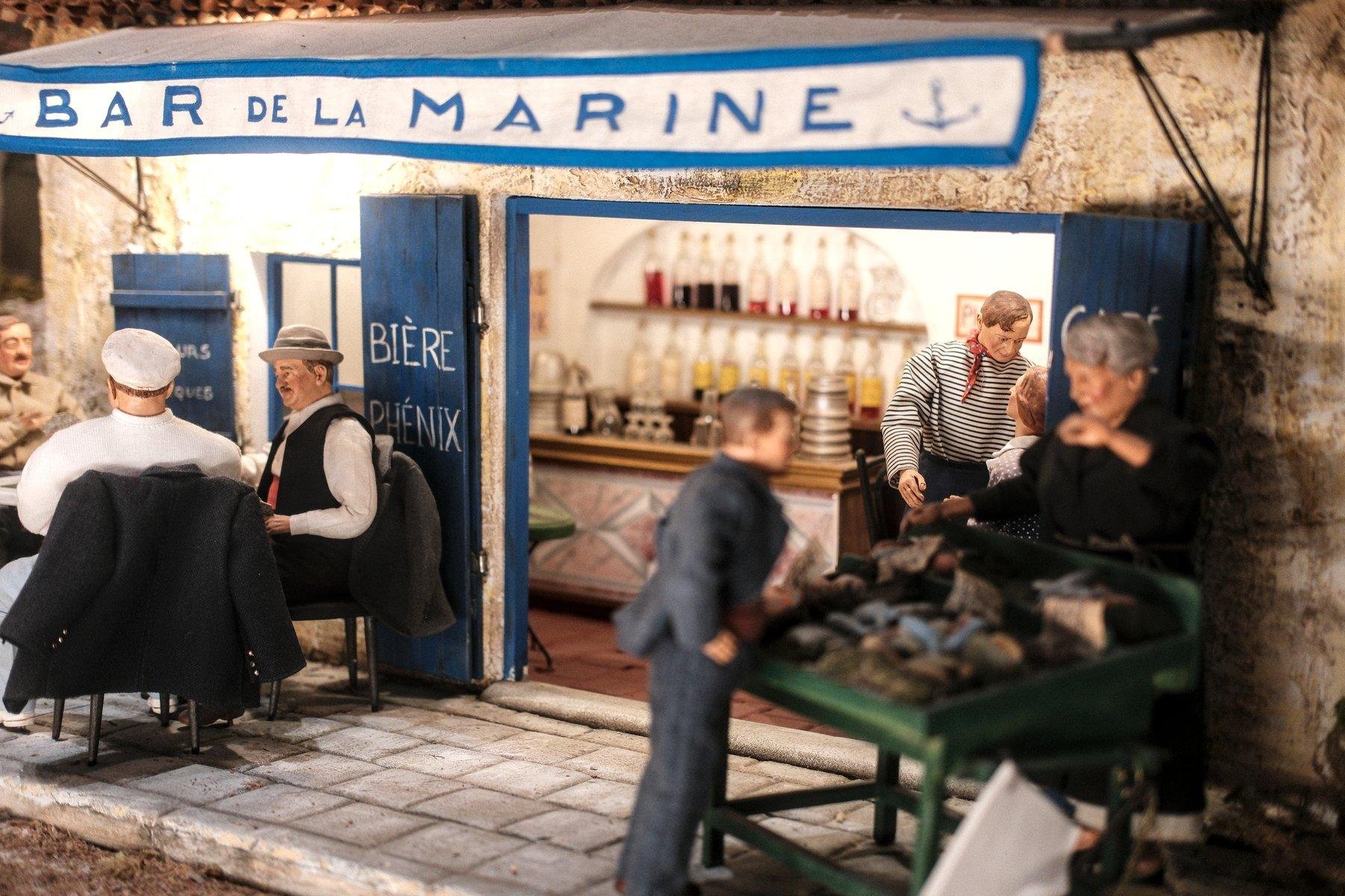 Photo horizontale. Gros plan sur un diorama en santons, représentant un bar, avec son store marqué «&nbsp;bar de la Marine&nbsp;», sa terrasse avec une table et des convives, un homme en marinière dans le bâtiment parlant à une femme. Il y a aussi un comptoir et derrière, des bouteilles sur étagères. Sur la droite, un stand où une dame vend des huîtres à un homme