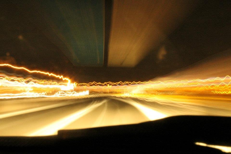 Foto en horizontal tomada desde el interior de un coche que está en movimiento en una autovía por la noche. Se aprecian las estelas luminosas y vibrantes de farolas, faros de otros coches, y formas movidas de elementos del paisaje como señales de tráfico y líneas de la carretera. La luminosidad es amarillenta.