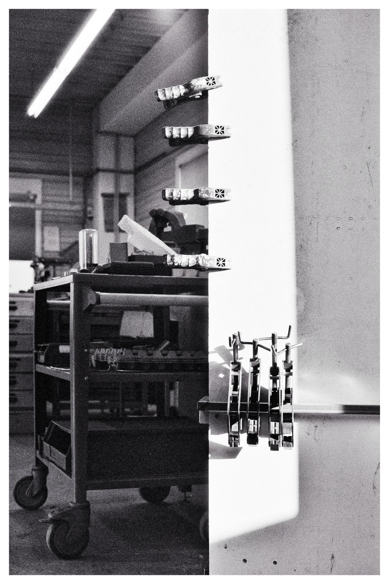 Clamps attached to a vertical white board on the right, casting shadows. To the left, a wheeled metal cart with tools and a glass sits in an industrial workspace with shelves and overhead fluorescent lighting. Black and white image with high contrast.