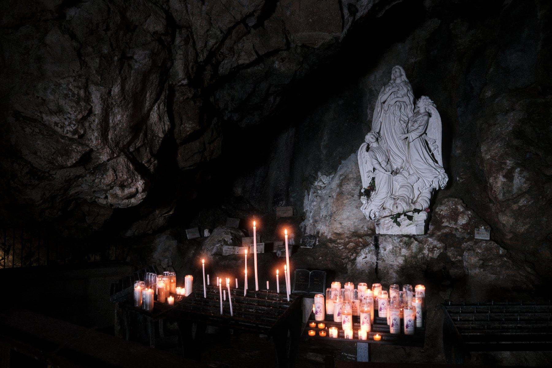 Photo horizontale. Dans une grotte sombre, une statue blanche de Marie-Madelaine, entourée de 2 anges sort des ténèbres. Des cierges brillent à ses pieds