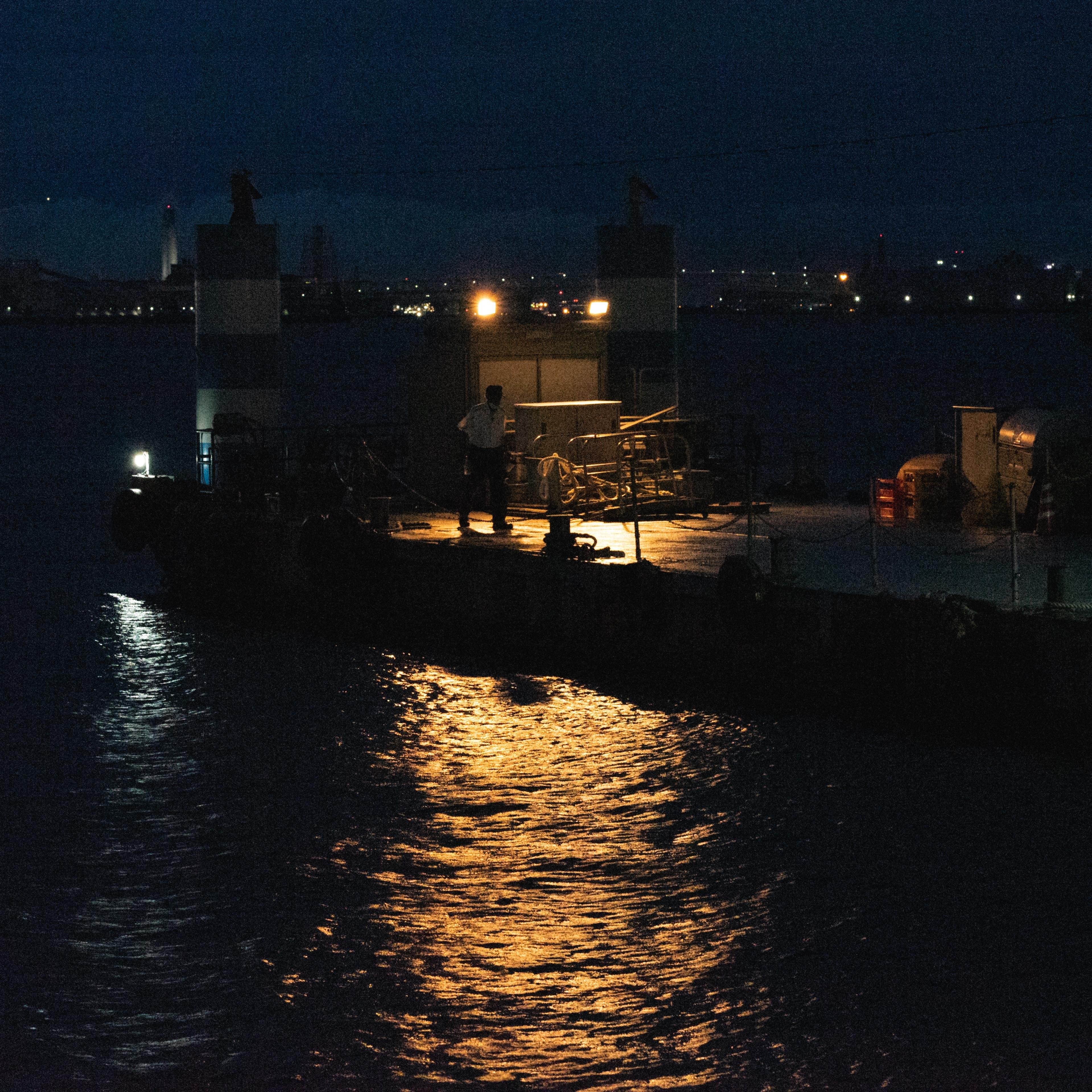 A person stands on a dimly lit dock at night, surrounded by equipment and barriers. Two bright lights illuminate the area, reflecting on the surface of the surrounding dark water. City lights are visible in the distant background under a dark sky.