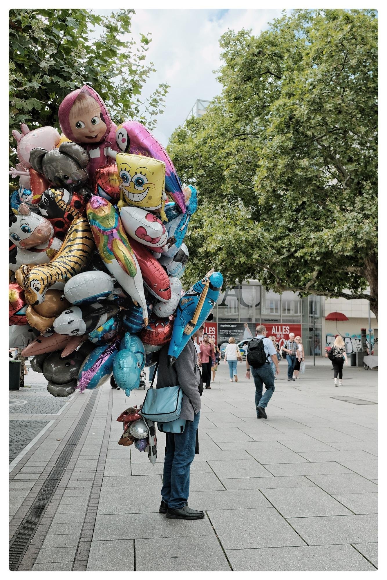 A person holding a large bundle of colorful, cartoon-themed helium balloons, including characters like SpongeBob SquarePants and Masha from Masha and the Bear. The person is wearing a gray jacket and blue jeans, partially obscured by the balloons. Background features a public square with several pedestrians walking, and large trees to the side.