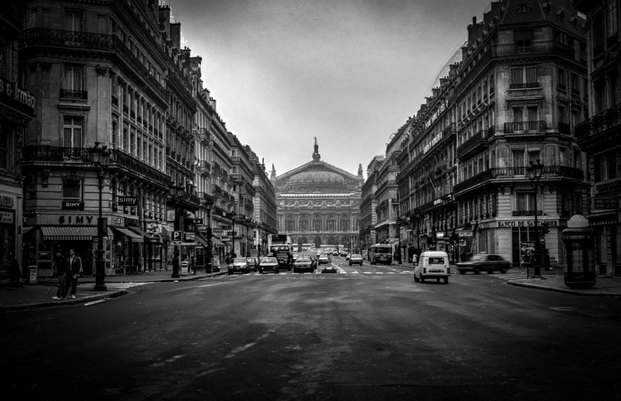 Wide city street flanked by tall, ornate buildings in a symmetrical composition. In the distance, a large domed building stands prominently. Cars are parked along the street, and a few vehicles and pedestrians are visible. The scene is in black and white, giving a classic, timeless atmosphere.