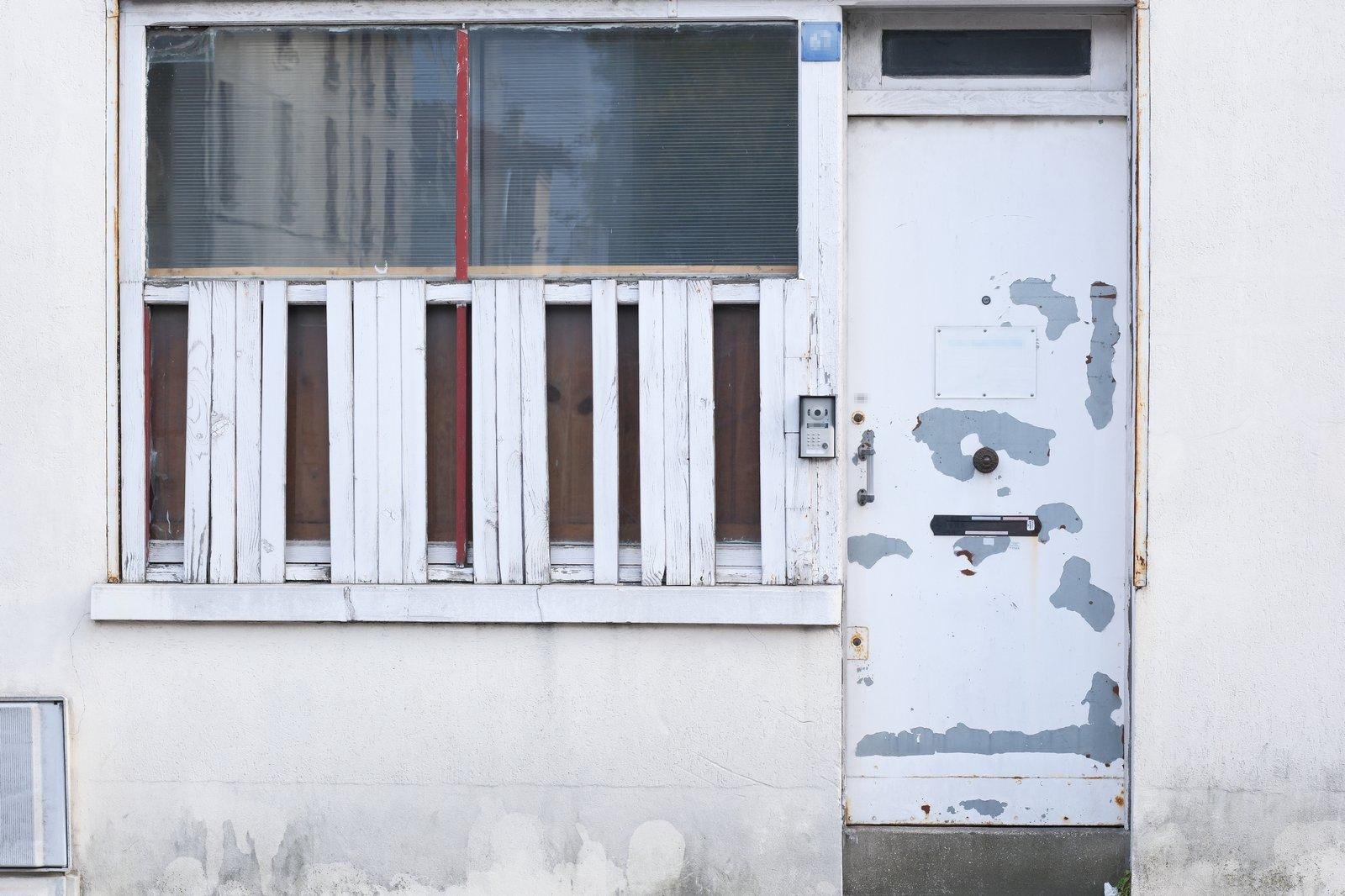 Photo format paysage, montrant une porte blanche à la peinture fortement écaillée dont des plaques entières sont tombées, laissant apparaître du gris. À gauche, une fenêtre dont la moitié basse est barricadée de planches blanches, mais il en manque pas mal. Une barre rouge barre la fenêtre verticalement au centre