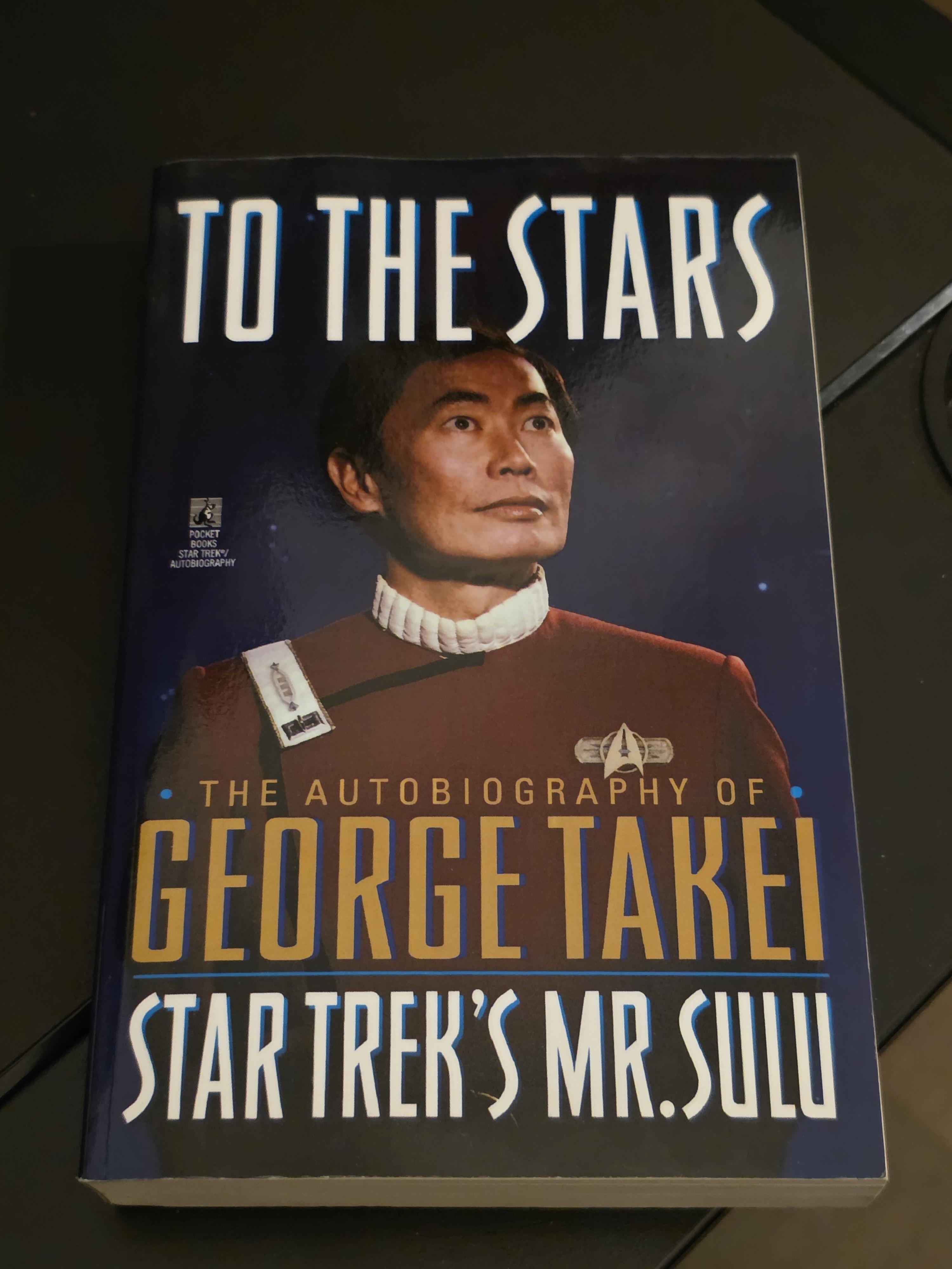 A paperback copy of "To the Stars," the autobiography of George Takei, rests on a dark surface. The cover features a heroic portrait of Takei looking toward the heavens, dressed in the classic maroon Starfleet uniform from the Star Trek film era, complete with a white turtleneck and silver insignia.
​The title is written in large, sleek white letters at the top, while Takei's name stands out in bold gold text across the center. Below that, the text "STAR TREK'S MR. SULU" is written in a stylized white font. Published in 1994, this memoir offers an intimate look at Takei's life, from his childhood spent in Japanese American internment camps during World War II to his groundbreaking career in Hollywood and his later work as a social justice activist. The background of the cover is a deep indigo cosmos filled with tiny, glimmering stars, reflecting the "Star Trek" spirit of exploration and hope.