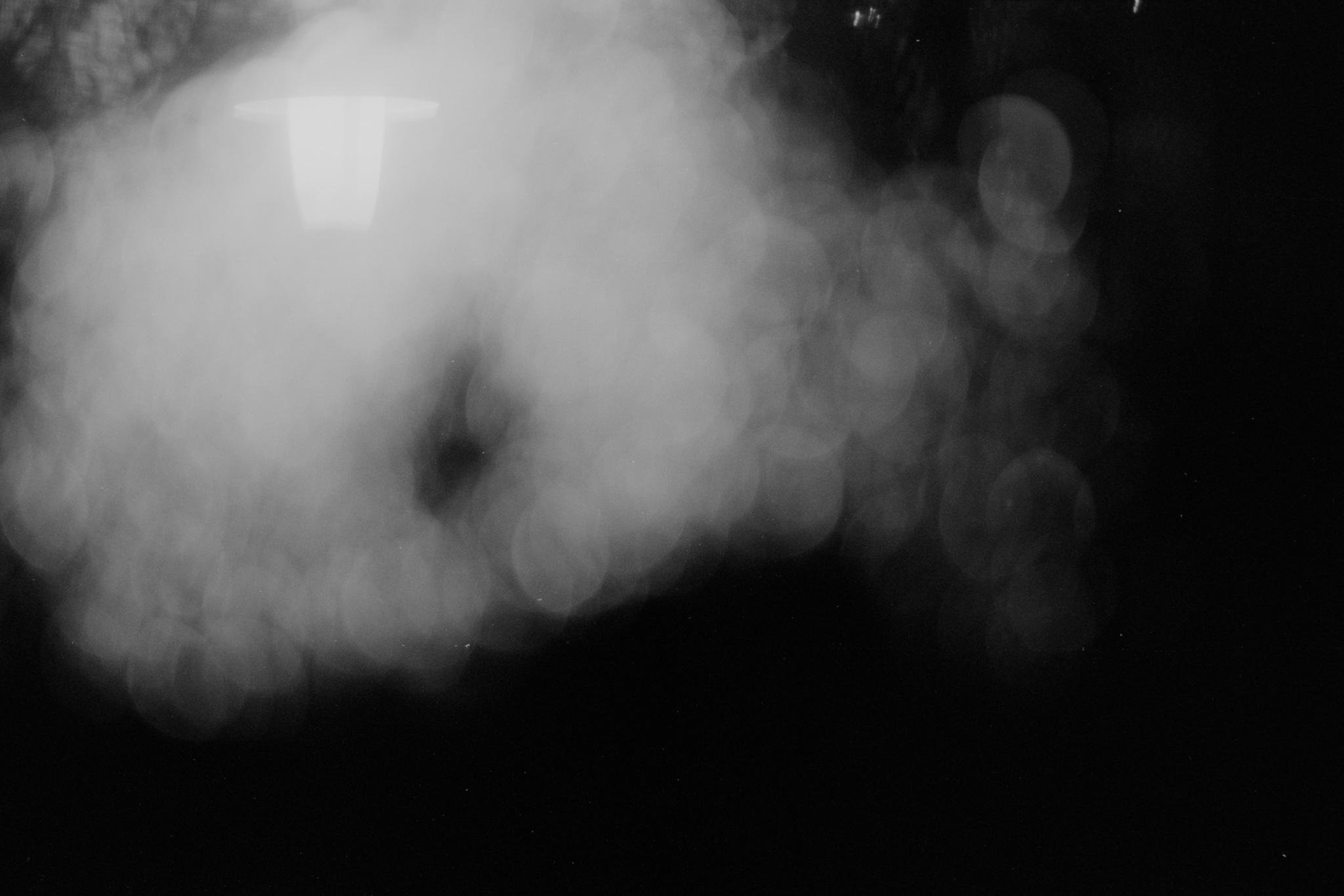 Blurred black-and-white photo with a large white hazy shape on the left and scattered circular light bokeh on the right against a dark background.