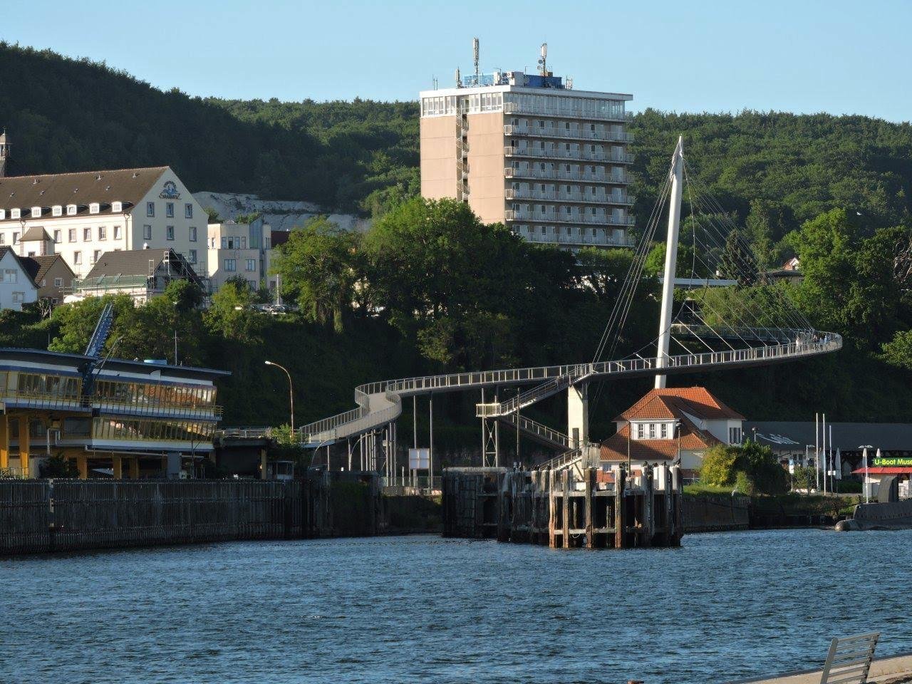 Curved pedestrian bridge with metal cables spans over a body of water. Several buildings, including a tall, beige apartment block and smaller structures with red roofs, are situated on a tree-covered hillside. The scene is set on a sunny day with clear skies.