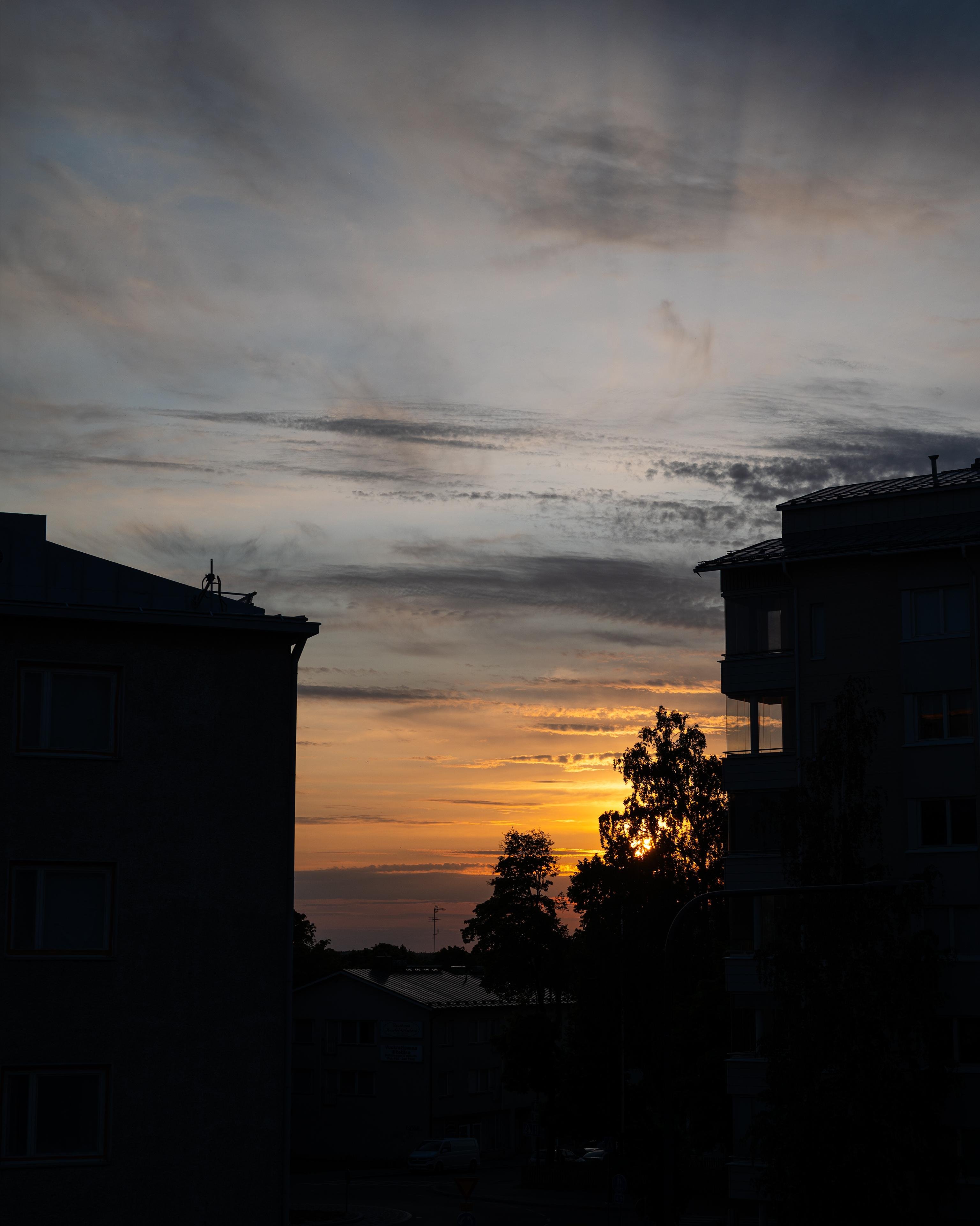 Sunset with an orange and yellow sky, partially obscured by wispy clouds. Silhouetted buildings frame the scene on either side, and tree outlines are visible in the foreground.