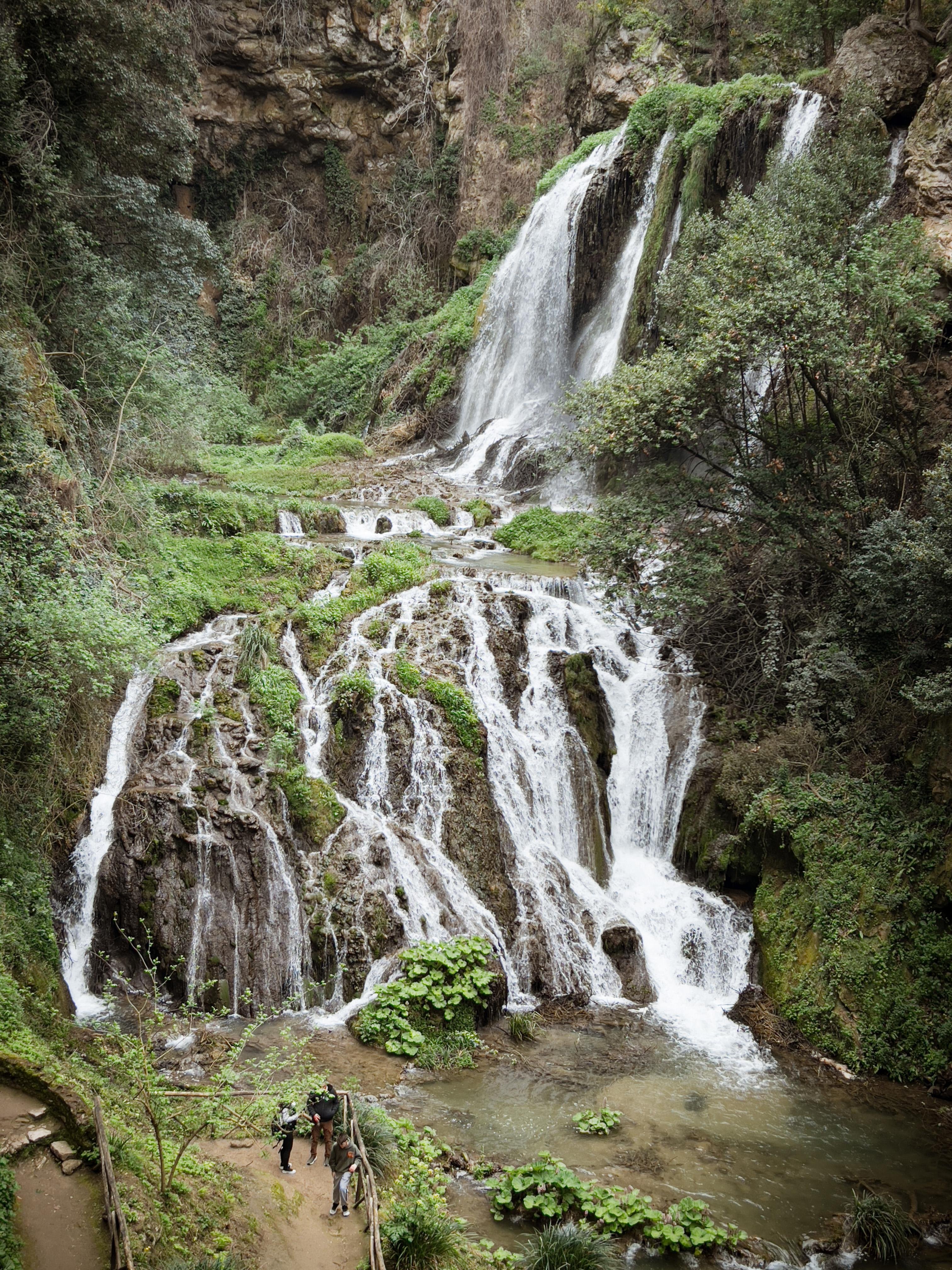 Vista panoramica di una cascata che scende da una scogliera rocciosa circondata da una rigogliosa vegetazione. Ci sono persone alla base della cascata, che aggiungono una scala alla scena. L'area sembra un ambiente naturale e sereno, ideale per escursioni o passeggiate nella natura.