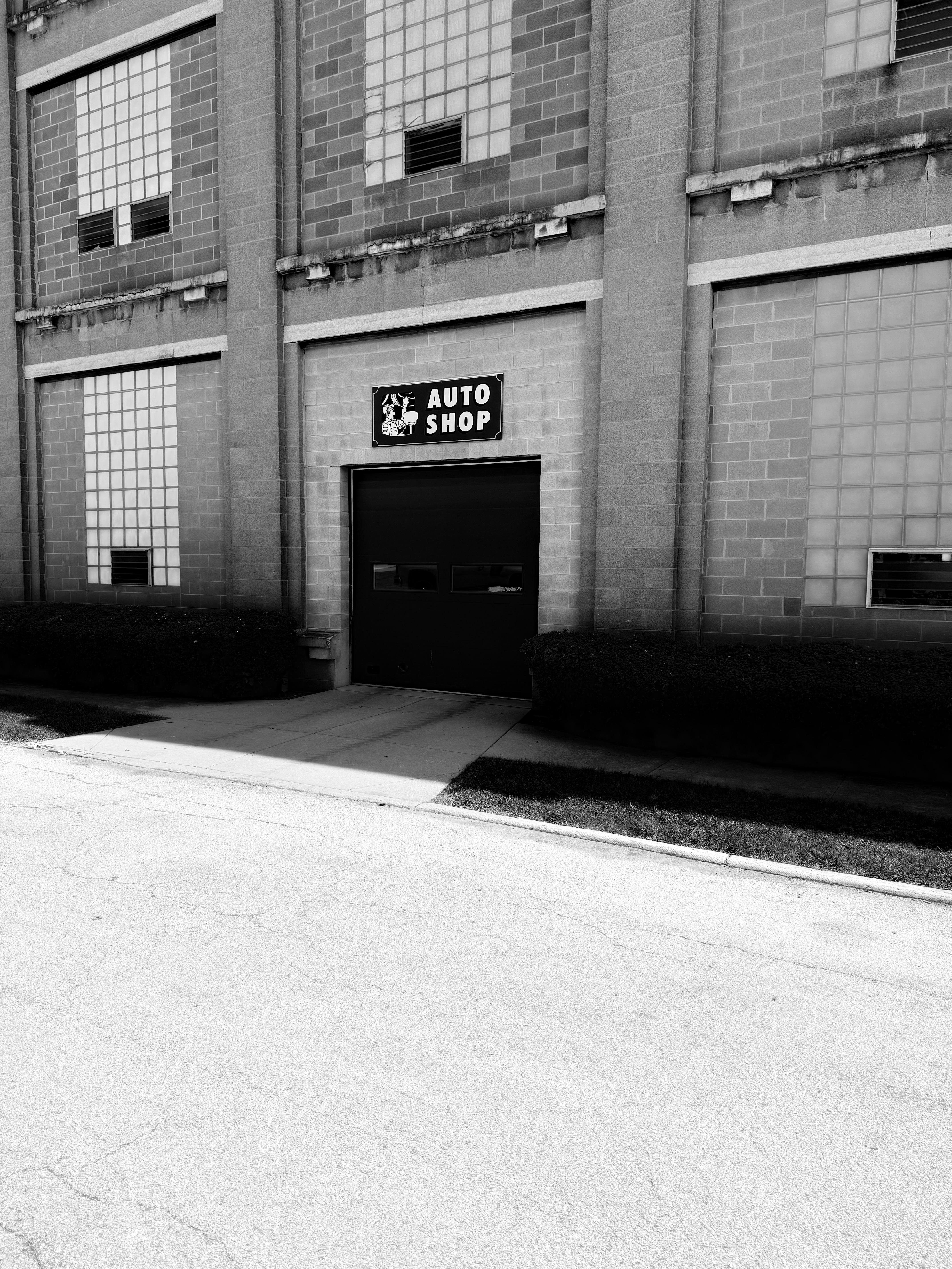 The image shows the exterior of a building with a sign that reads "AUTO SHOP." The structure has large glass block windows and features typical industrial architecture. There are shrubs lining the path leading to the door.