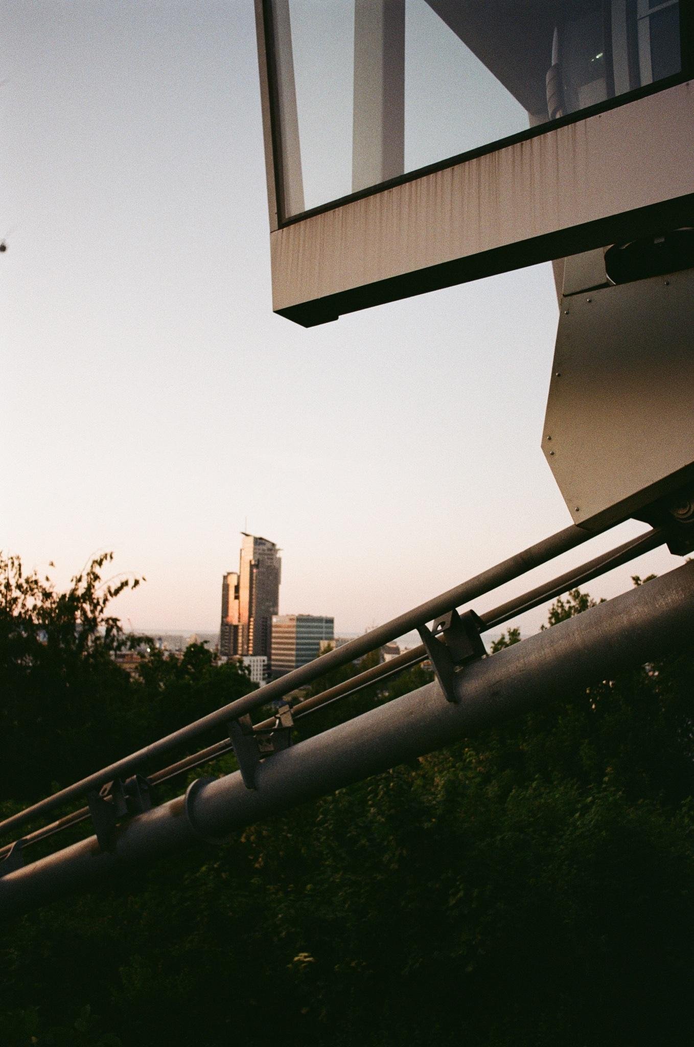 Diagonal metal support beams and part of a glass-and-metal structure in the foreground, with treetops below and a distant city skyline of tall buildings against a pale sunset sky.