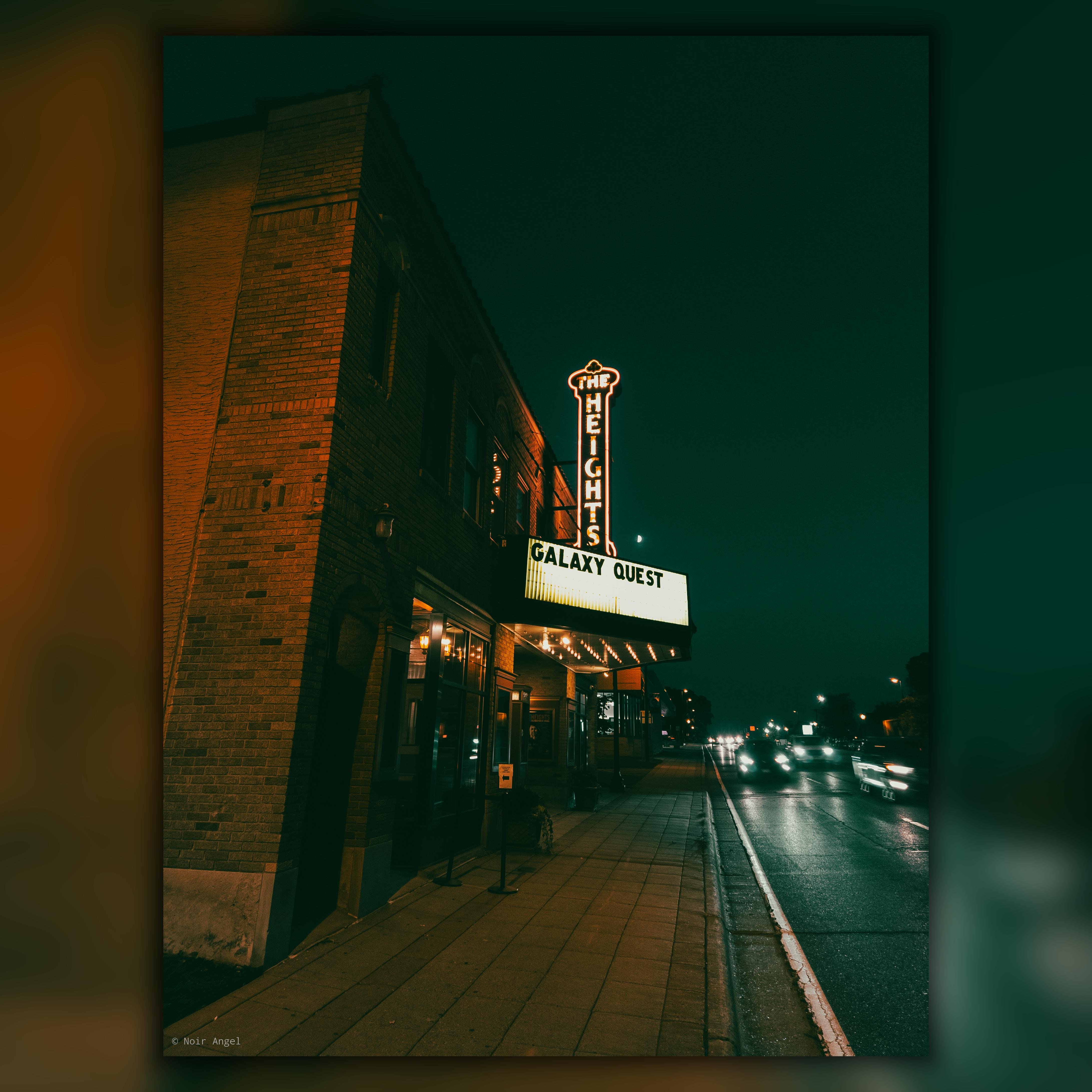 Brick theater building on a street corner at night, illuminated by a neon sign reading "THE HEIGHTS" and a marquee displaying "GALAXY QUEST." The sidewalk is empty, and car lights streak by on the adjacent road, creating a sense of motion in the dark environment.