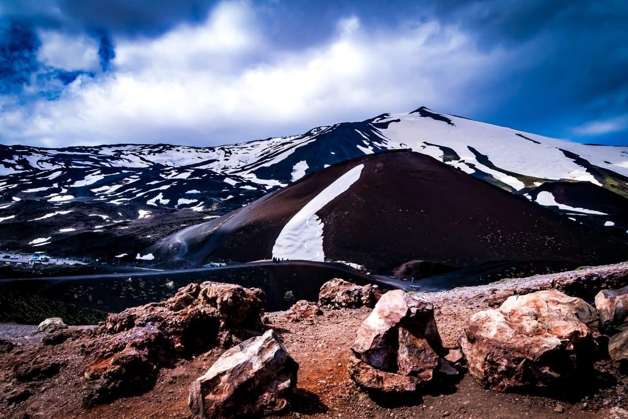 Rocky foreground with large boulders overlooking dark volcanic hills streaked with snow, with snow-capped mountains in the background under a cloudy blue sky.