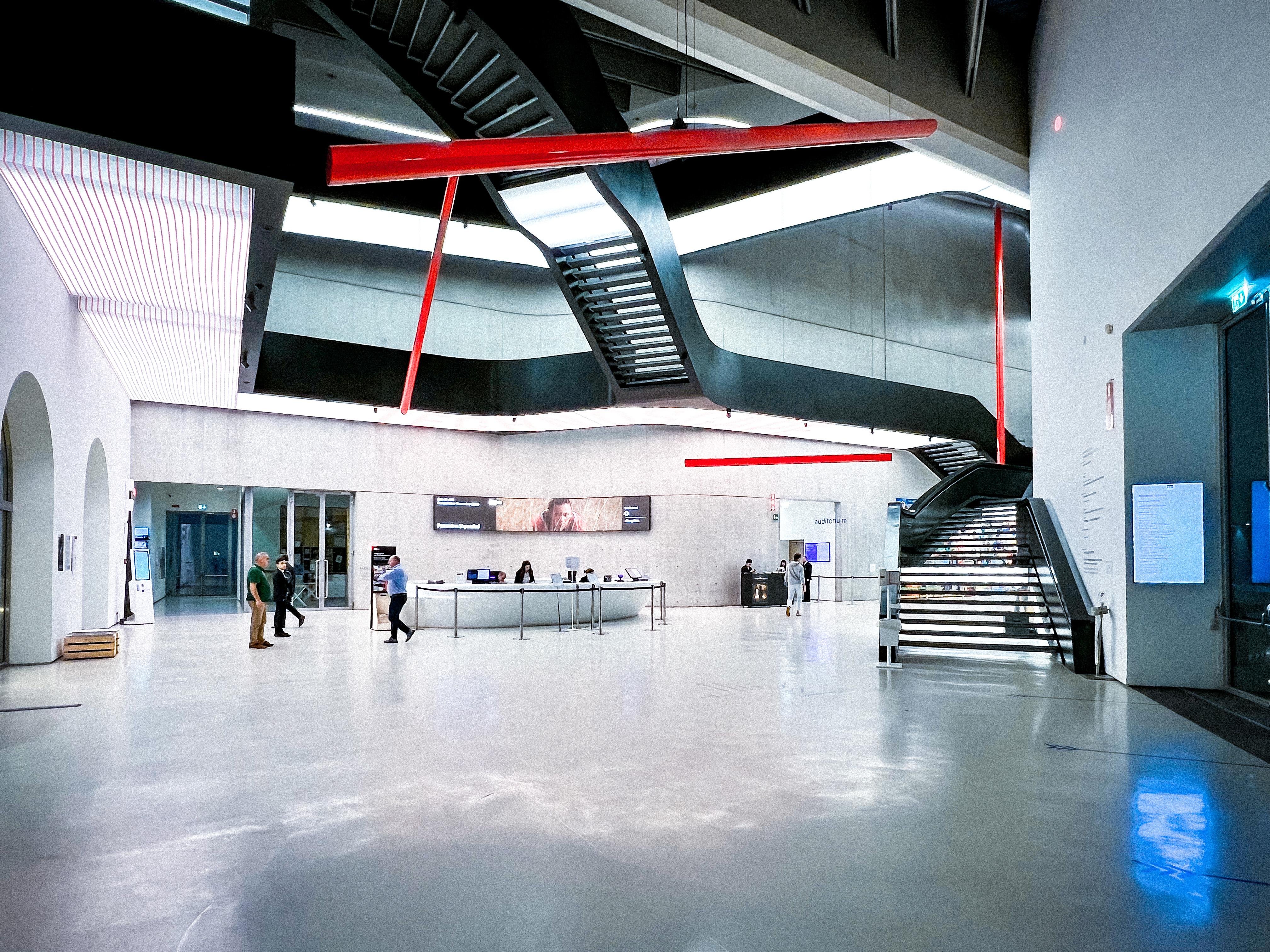Interno di un edificio moderno con un'area spaziosa e aperta, caratterizzato da un'architettura elegante. Al centro c'è una grande reception, alcune persone che camminano e scale nere con accenti rossi ben visibili. Il design è minimalista con linee pulite e un focus sulle forme geometriche. L'illuminazione è brillante, migliorando l'estetica contemporanea.