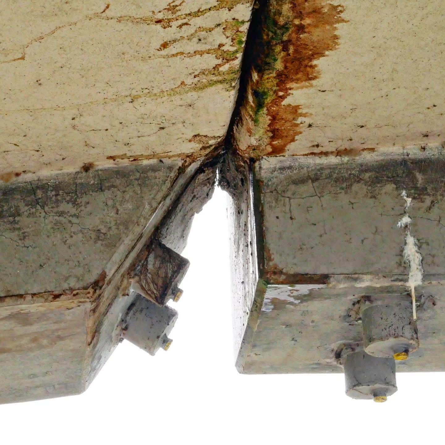 This image shows a structural joint or connection between two concrete sections, part of a bridge. The surfaces display signs of wear, such as rust stains, which might indicate aging. There are bolts visible, likely used to secure the connection.
