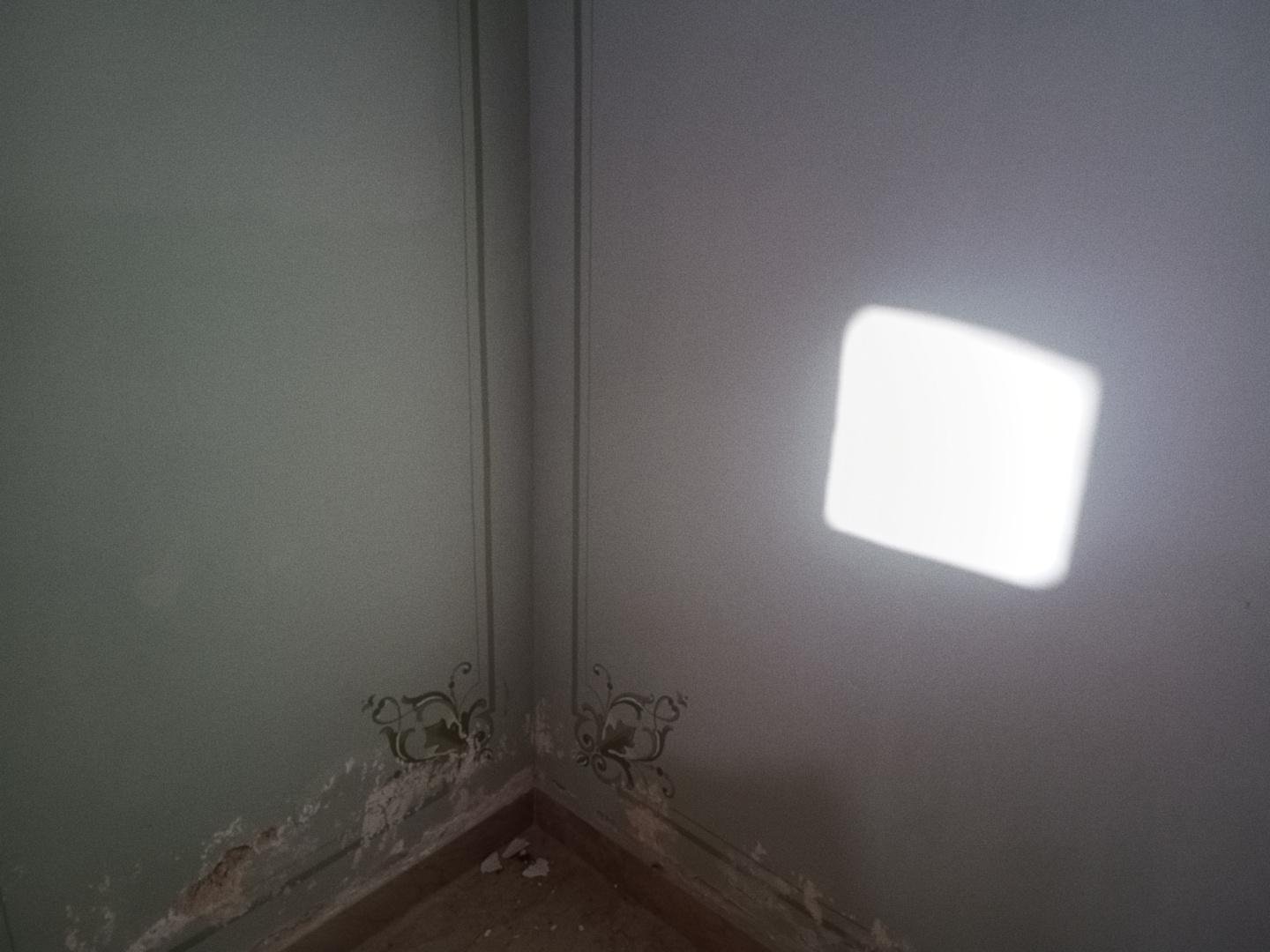 Angolo screpolato tra due muri, decorato con motivi floreali. Sulla parete a destra è visibile un quadrato di luce filtrata da un'apertura sul tetto, fuori inquadratura.