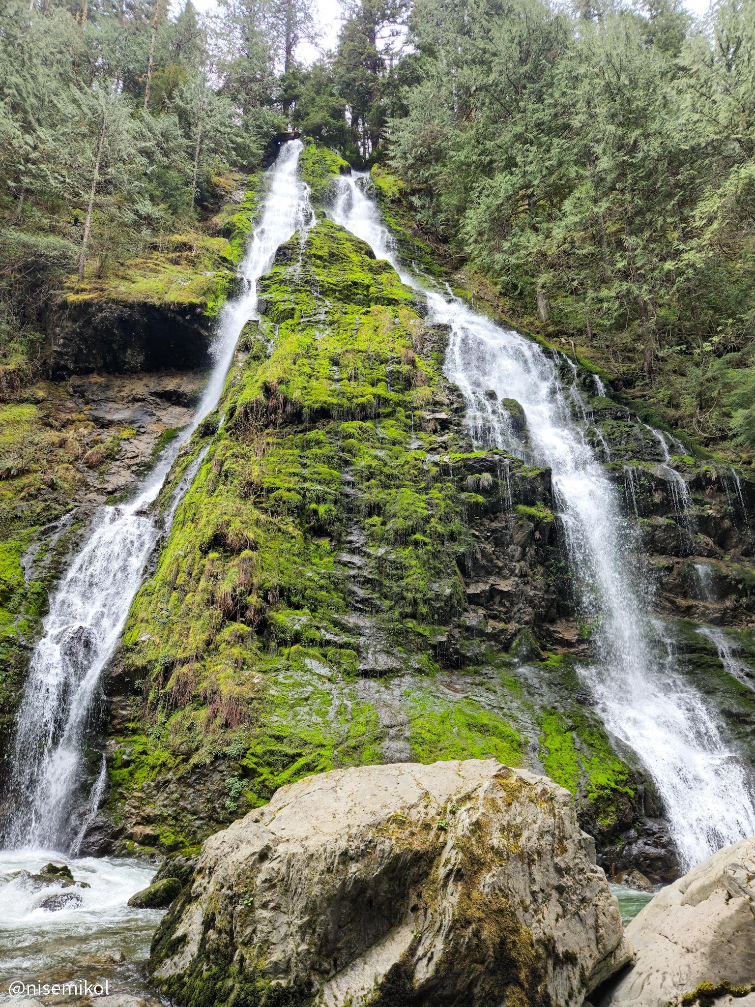 Waterfall cascading down a moss-covered rocky cliff surrounded by lush green trees. The water splits into two streams as it flows over the bright green moss, converging into a pool at the base. Large rocks are visible in the foreground, and the surrounding area is forested with a mix of tall trees.