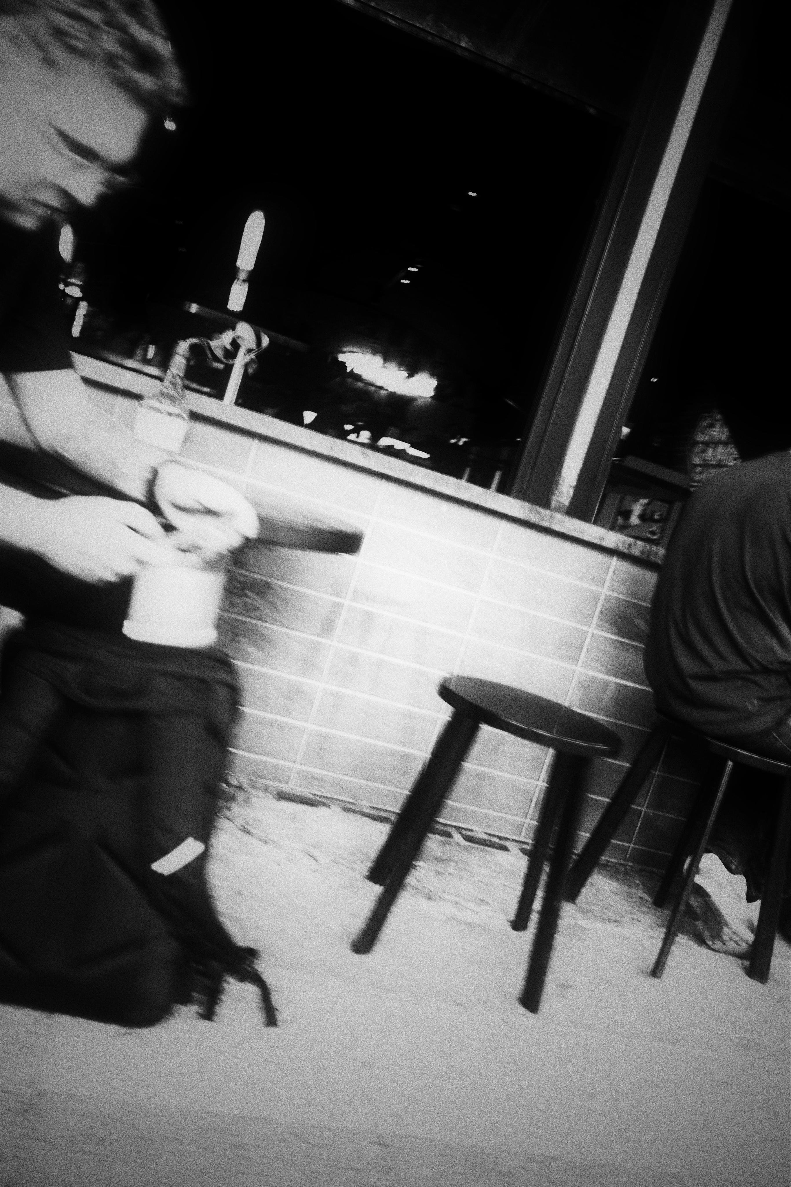 A person sitting on a stool looks at a phone. Their backpack is in front of them. Another person sits on a stool, back to the camera. An empty stool sits between them and an open window is behind them showing darkness inside.
