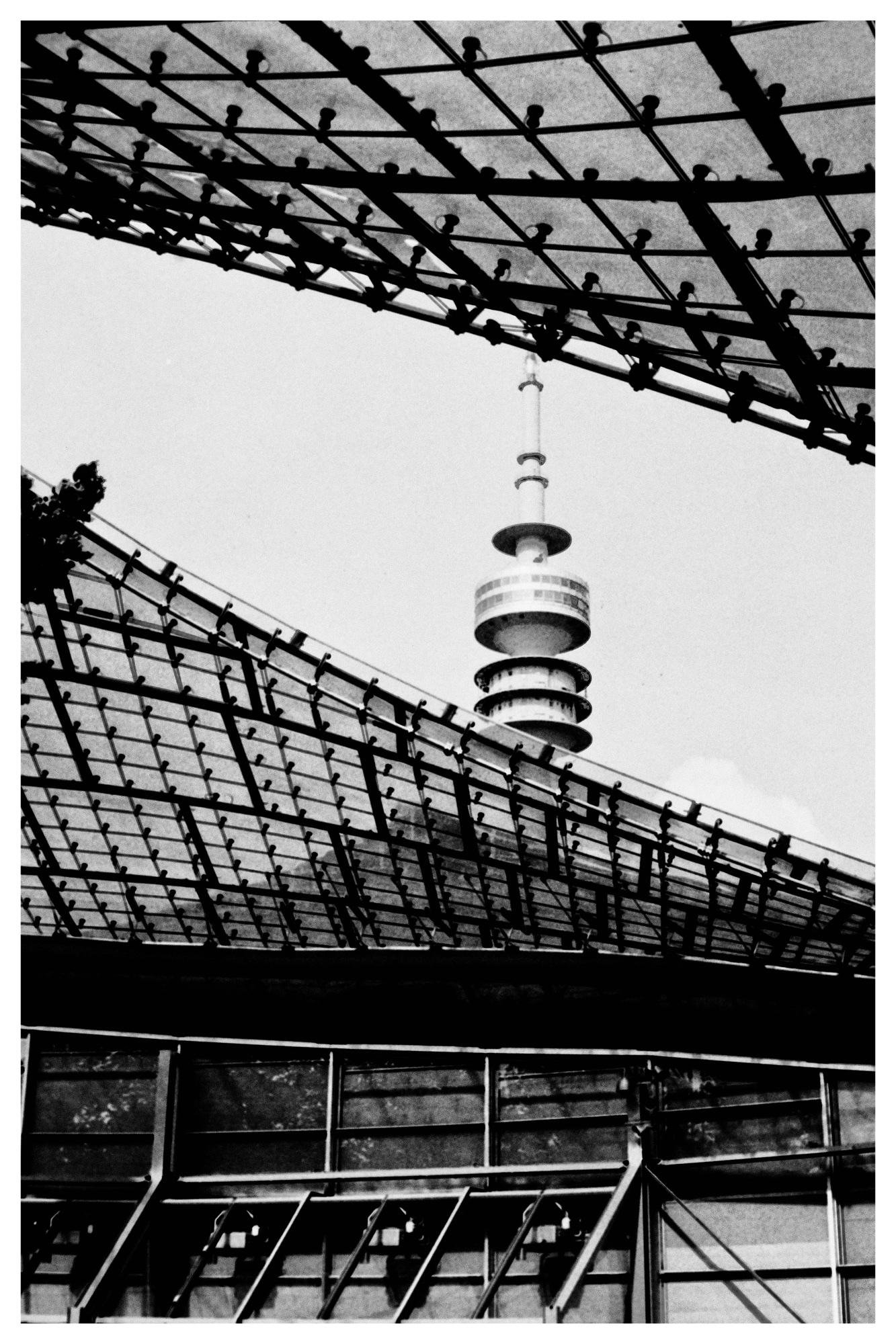Black-and-white upward view of a tower with stacked circular platforms and a thin antenna, framed by two large curved grid-like roof structures, with a section of building facade along the bottom.